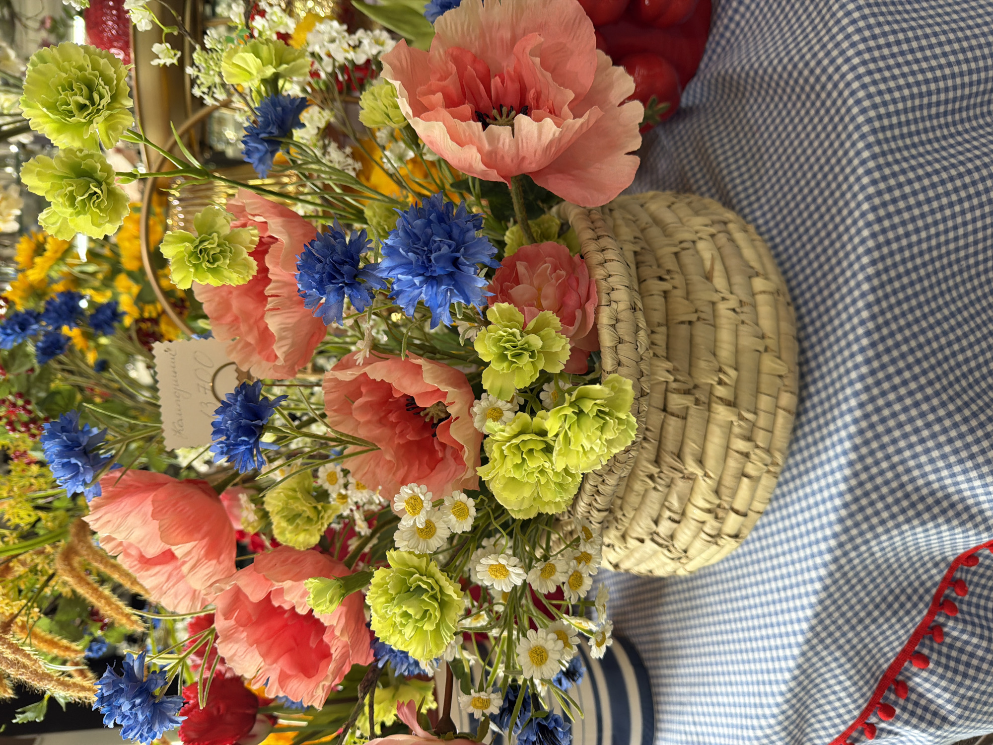 The image shows a still life arrangement featuring a woven basket overflowing with an assortment of flowers. The flowers include pink poppies, blue cornflowers, small white daisies, and lime green carnations. The arrangement is placed on a blue and white checkered tablecloth with a red pom-pom trim. A blue and white striped plate is partially visible beneath the basket. The image does not provide enough information to determine if this image was taken in the city of Votkinsk, Russia. The location of the image can not be confirmed.