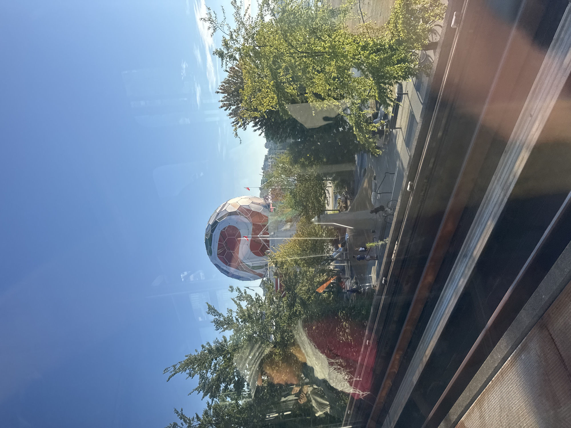The image shows a view of Science World at TELUS World of Science in Vancouver, Canada. The iconic geodesic dome, painted in red and white geometric patterns, is visible in the background under a clear blue sky. Trees with green foliage surround the building, and the foreground features what appears to be a train or tram track, with metal rails reflecting the sunlight. Several people are seen walking in the outdoor area around Science World, with some sitting at benches and tables. The image captures a bright, sunny day with clear visibility.