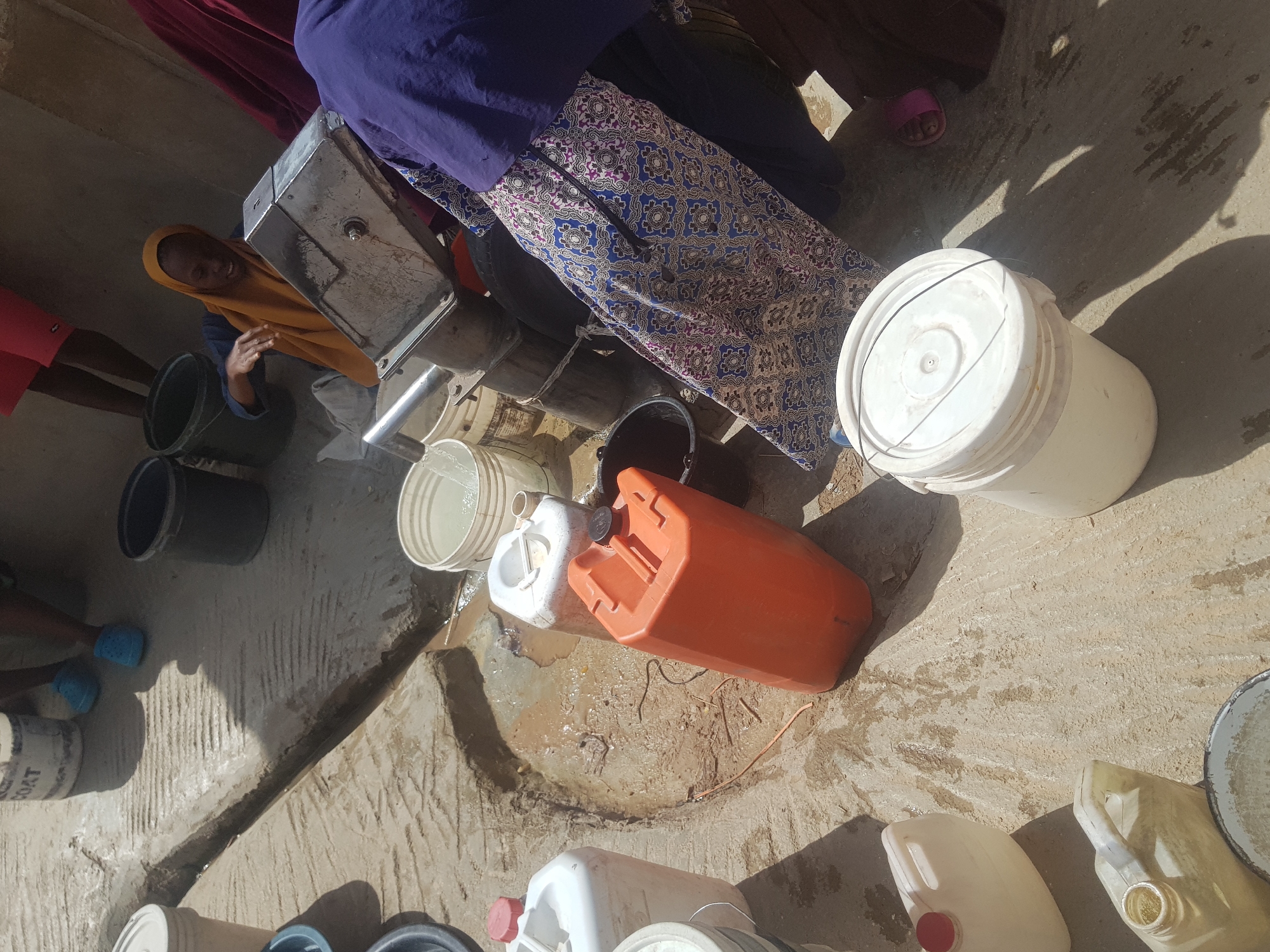 The image depicts a water distribution point in Dutsinma B, Nigeria. A manual pump stands on a concrete platform. Several people, including a woman in a yellow hijab, are gathering water. Numerous buckets and jerrycans, some full, surround the pump. The area is wet and muddy.