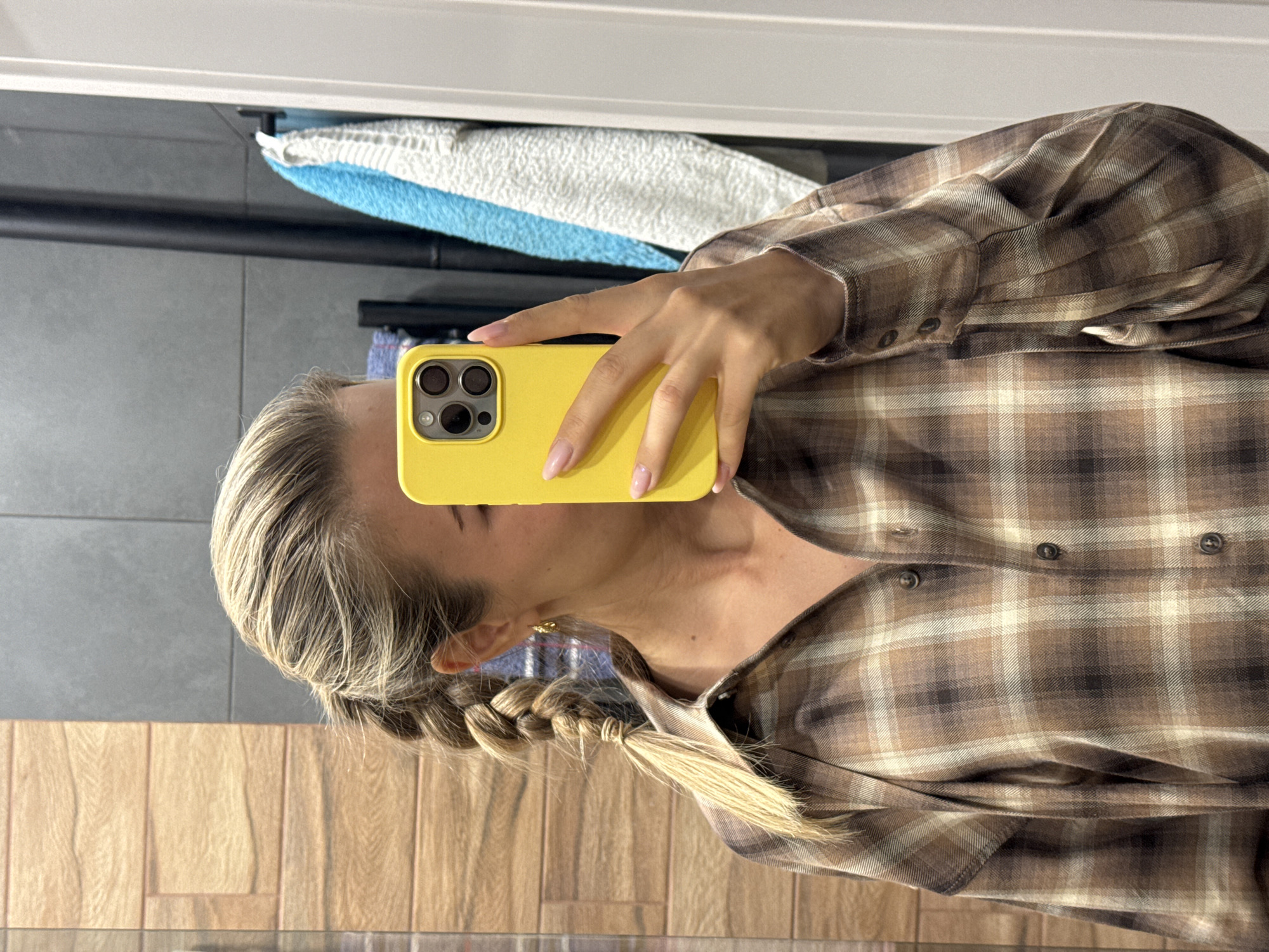 A woman with blonde braided hair is taking a selfie in a bathroom. She is wearing a brown and white plaid shirt. She is holding a yellow smartphone in front of her face. Behind her, there is a mirror, a grey tile wall, a wood-paneled wall, and a white and blue towel hanging on a black rod. The location is Unknown, Unknown.