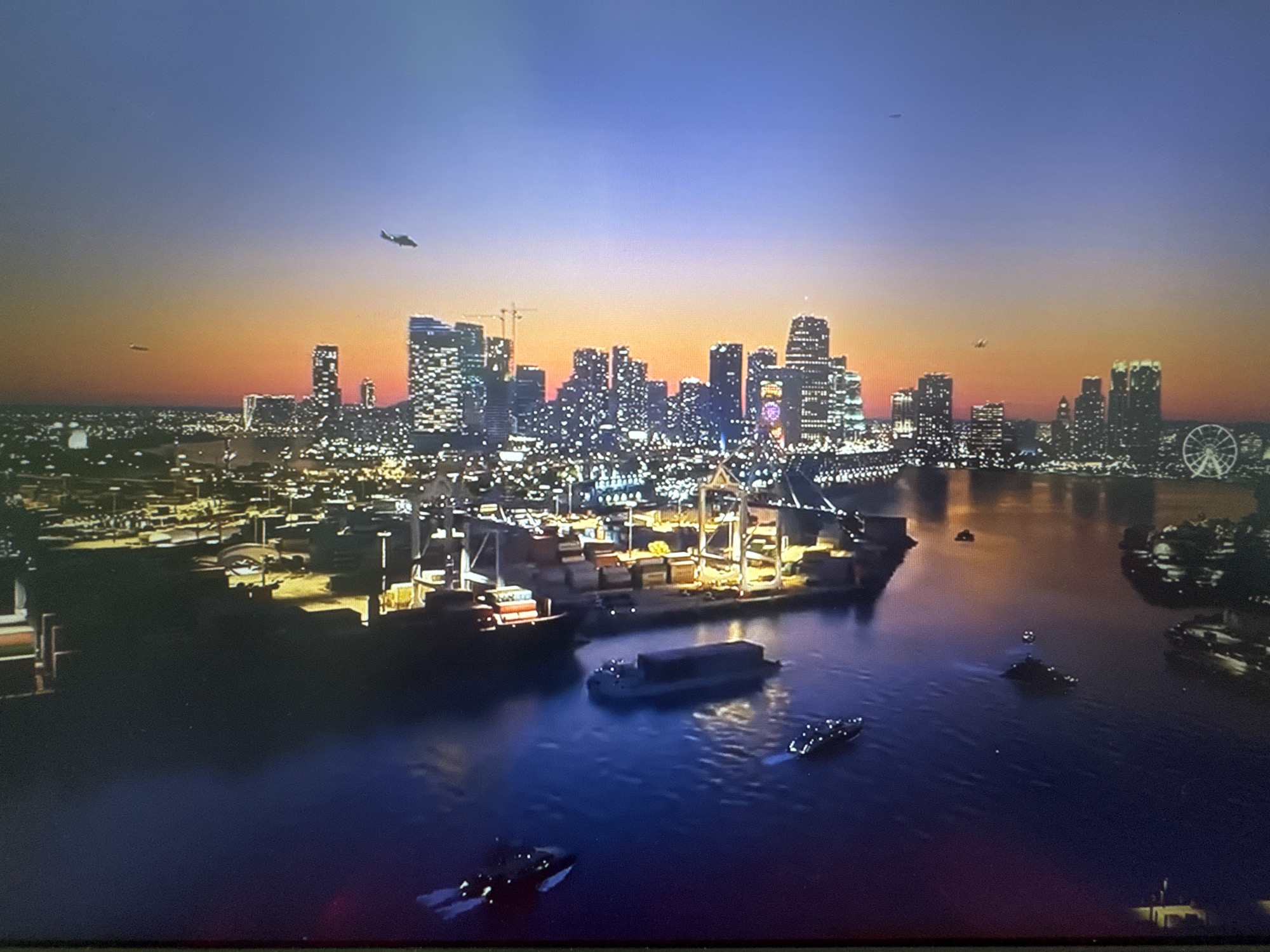 This is a night-time aerial view of a harbor and city skyline at dusk. The sky transitions from a deep blue at the top to orange and yellow near the horizon. The city features numerous illuminated skyscrapers. In the harbor, several cargo ships and smaller vessels are visible. One large cargo ship is docked at a loading crane facility with many shipping containers. The water reflects the lights of the city and the vessels. There are two airplanes visible in the sky. The image does not depict the city administration of Stepnogorsk, Kazakhstan, but rather a large coastal city.