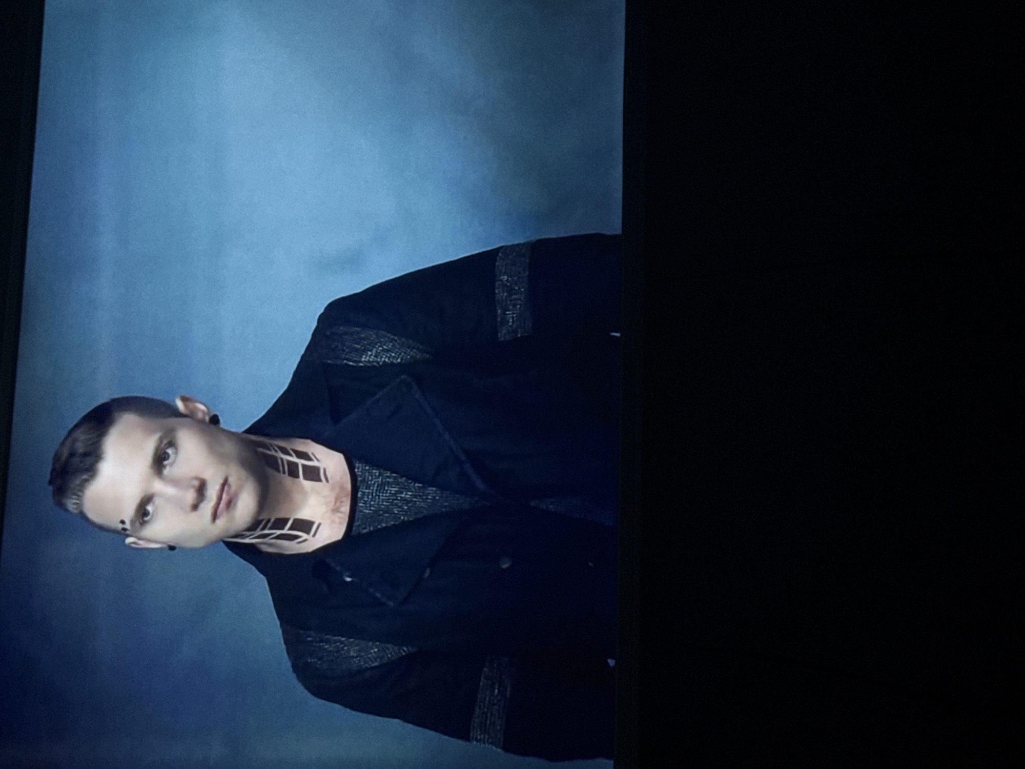 A man with light skin and short brown hair, wearing a black jacket and a gray shirt, is positioned against a mottled blue background. He has dark markings on his forehead and geometric tattoos on his neck. He appears to be facing the camera with a neutral expression. The right side of the image is black.