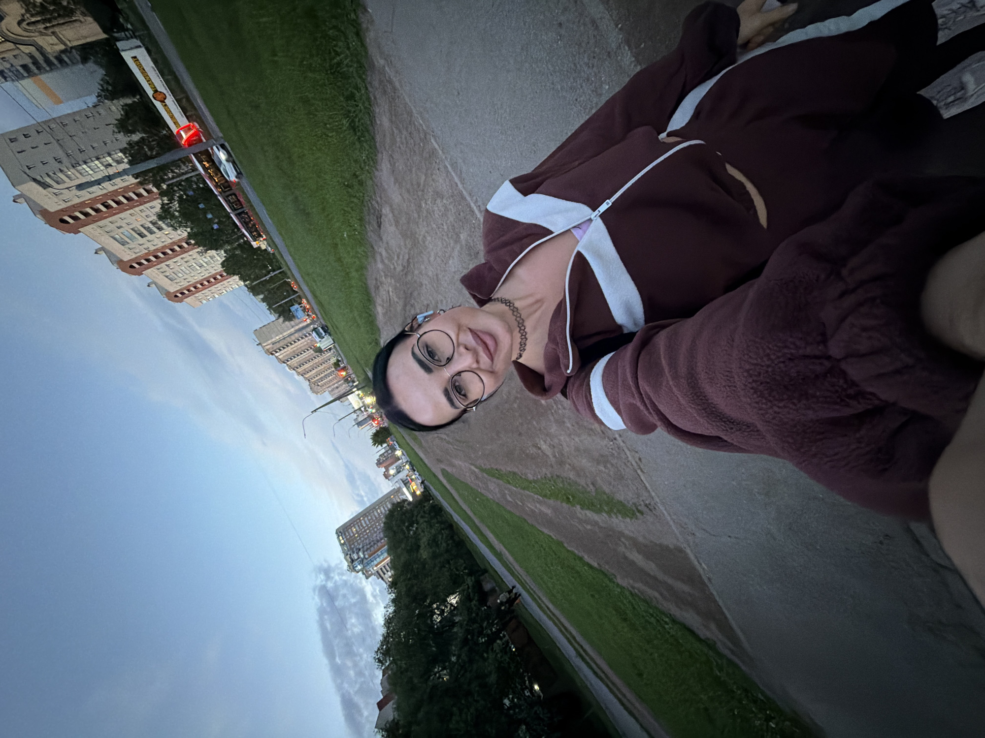 The image is a tilted selfie featuring a young adult positioned outdoors, likely in a park or along a street in Saint Petersburg, Russia, during dusk or early evening. The subject is a person with dark hair, wearing round glasses and a dark choker necklace, with earbuds in their ears. They are dressed in a brown hooded jacket with distinctive white stripes along the zipper and sleeves, and a light-colored top visible underneath. A faint smile is on their face as they look towards the camera.
The background reveals an urban setting with a paved path or road, a strip of green grass, and trees. Beyond these, multiple-story buildings with illuminated windows line the horizon, suggesting an active city environment. A white tram with red details is visible on tracks in the mid-ground; it prominently displays the "Coca-Cola" text and logo on its side. Streetlights are lit, and faint vehicle lights can be seen in the distance. The sky transitions from a lighter blue near the horizon to a deeper hue overhead, with scattered clouds, indicating the time of day. The overall atmosphere is tranquil, with the subject appearing calm and relaxed.