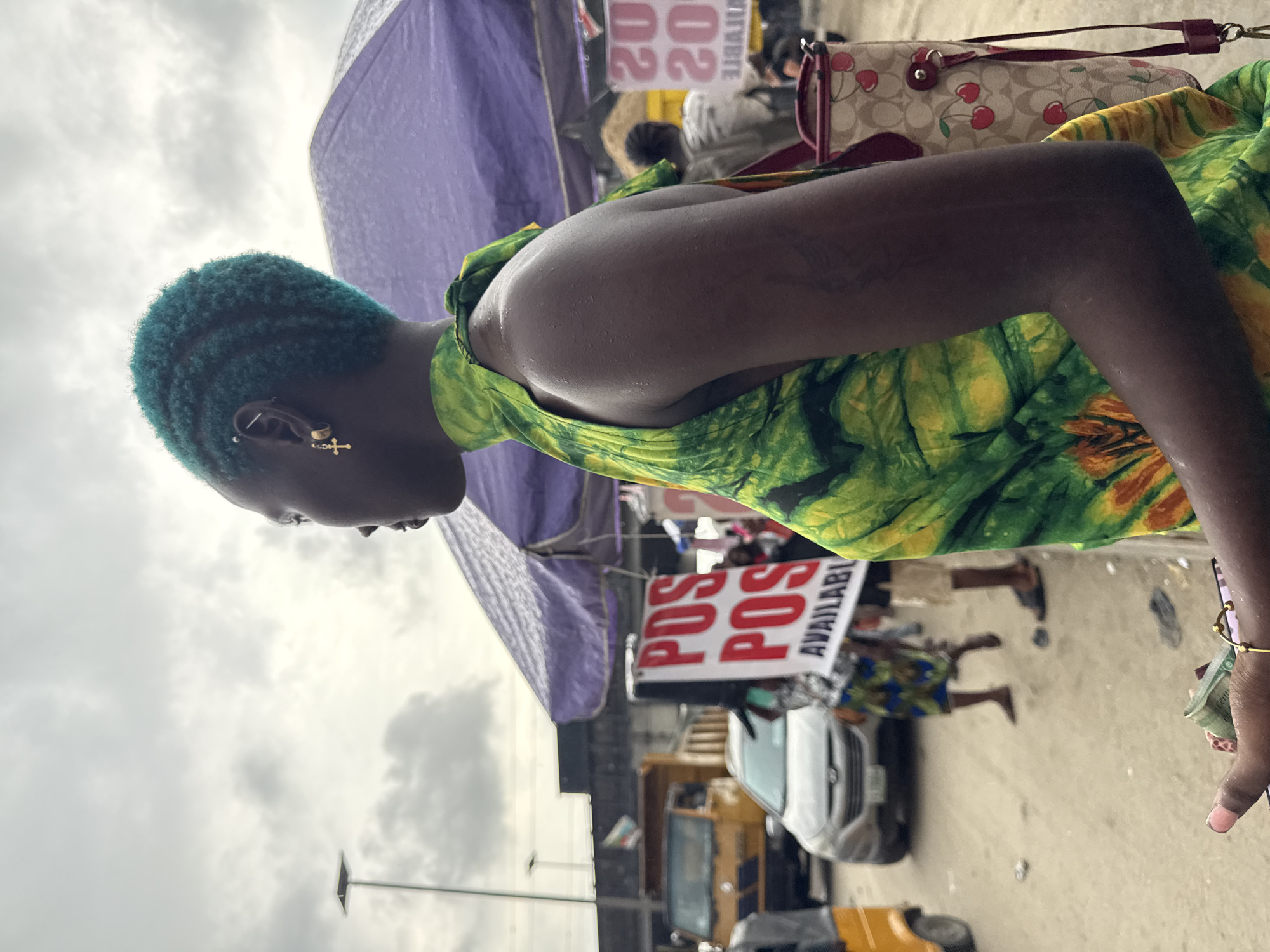 In Lekki, Nigeria, a woman with vibrant teal hair is seen from the side in a bustling street scene. She wears a bright green and yellow tie-dye dress and a gold cross earring. Her hand, adorned with a gold bracelet, holds a wad of Nigerian currency. In the background, a yellow truck, a silver car, and a three-wheeled vehicle are visible on the dirt road. Several stalls with purple awnings line the street, with signs indicating "POS AVAILABLE" and "20 20". Other individuals are present in the distance, suggesting a lively market environment. The sky is overcast, hinting at a humid or rainy day.