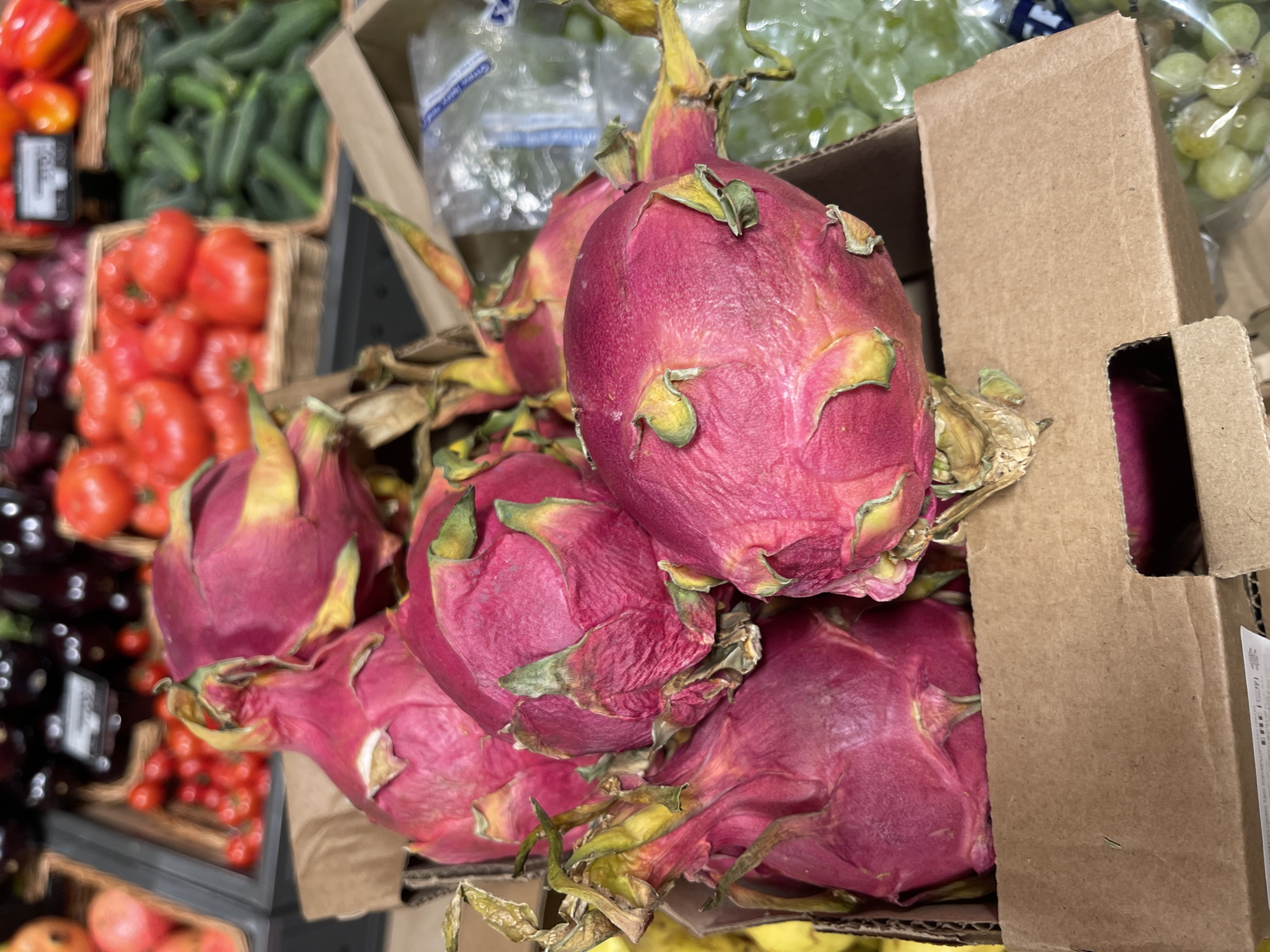 The image displays a vibrant selection of fresh produce, characteristic of a grocery store or market, possibly in Moscow, Russia. The central focus is a brown cardboard box overflowing with numerous dragon fruits (pitaya). These fruits are distinctly bright magenta-pink with green and yellow scales, some of which appear slightly withered.
In the blurred background on the left, various other fruits and vegetables are visible in baskets. Above, there's a quantity of green, elongated vegetables, resembling cucumbers. Below these, a basket is filled with red tomatoes, while further left, other red and darker-colored produce (possibly onions or eggplants) can be seen. An unreadable label or price tag is present among these items.
On the right side of the image, portions of the cardboard box are visible. Behind and above the dragon fruits, out of focus, are clear plastic bags containing green grapes. An illegible white label with black text and a barcode is attached to the side of the main cardboard box. The lighting suggests an indoor setting, likely under artificial lights. There are no people visible, and no specific time of day or weather can be determined from the image.