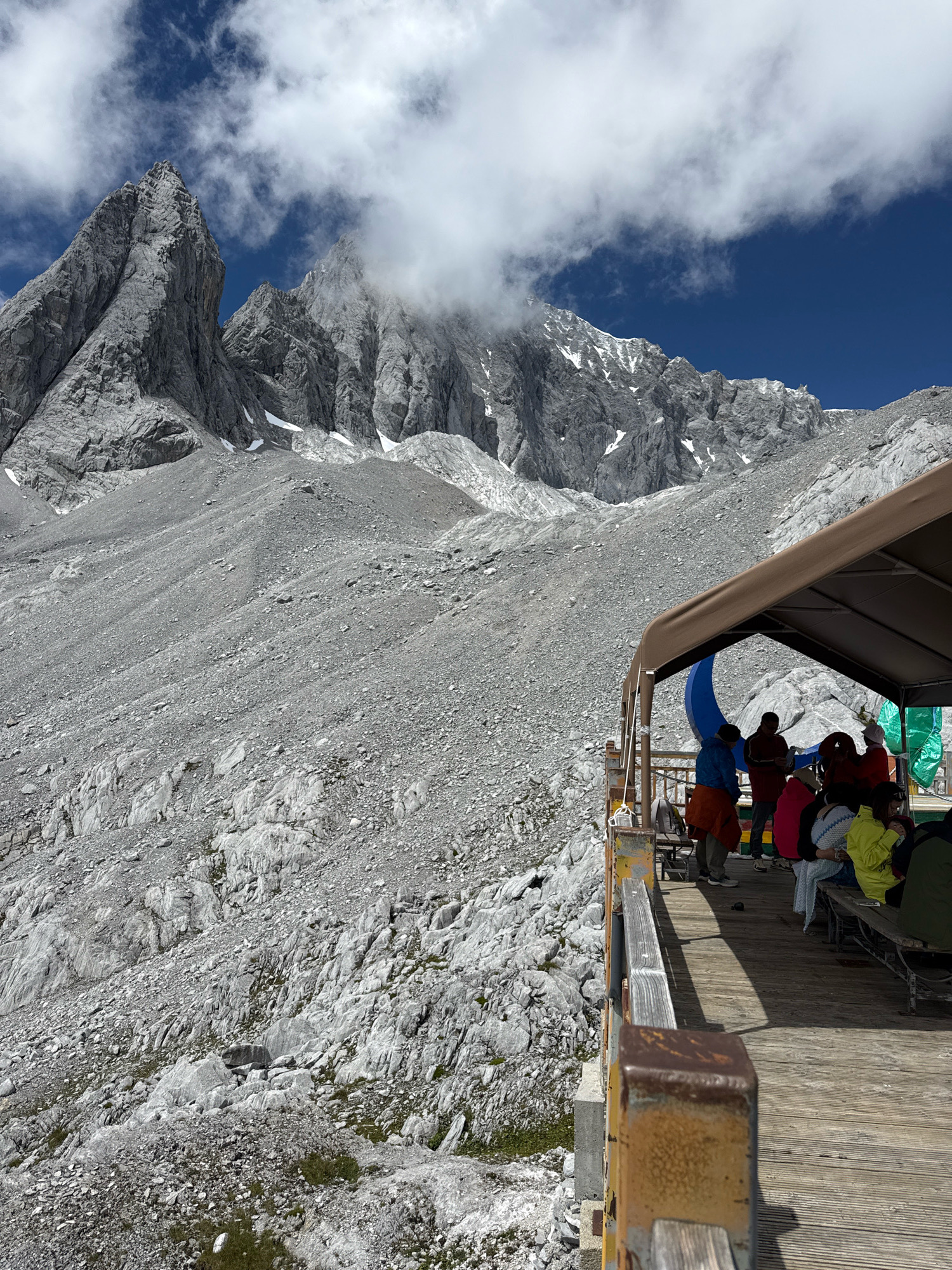 The image depicts a scenic mountain landscape in Yulong Nakhi Autonomous County, China, likely at a high altitude. In the foreground, a wooden platform with a brown awning provides shelter for a group of people seated on benches. The platform has a rustic metal railing that shows signs of age. The people are casually dressed, suggesting they are tourists. Behind the platform, the terrain consists of a vast, rocky, grey mountainside that stretches towards dramatic, jagged peaks in the background. The peaks are partially obscured by white clouds, and patches of snow are visible on their slopes. The sky is a vibrant blue. The environment appears rugged and natural, characteristic of a mountainous region.