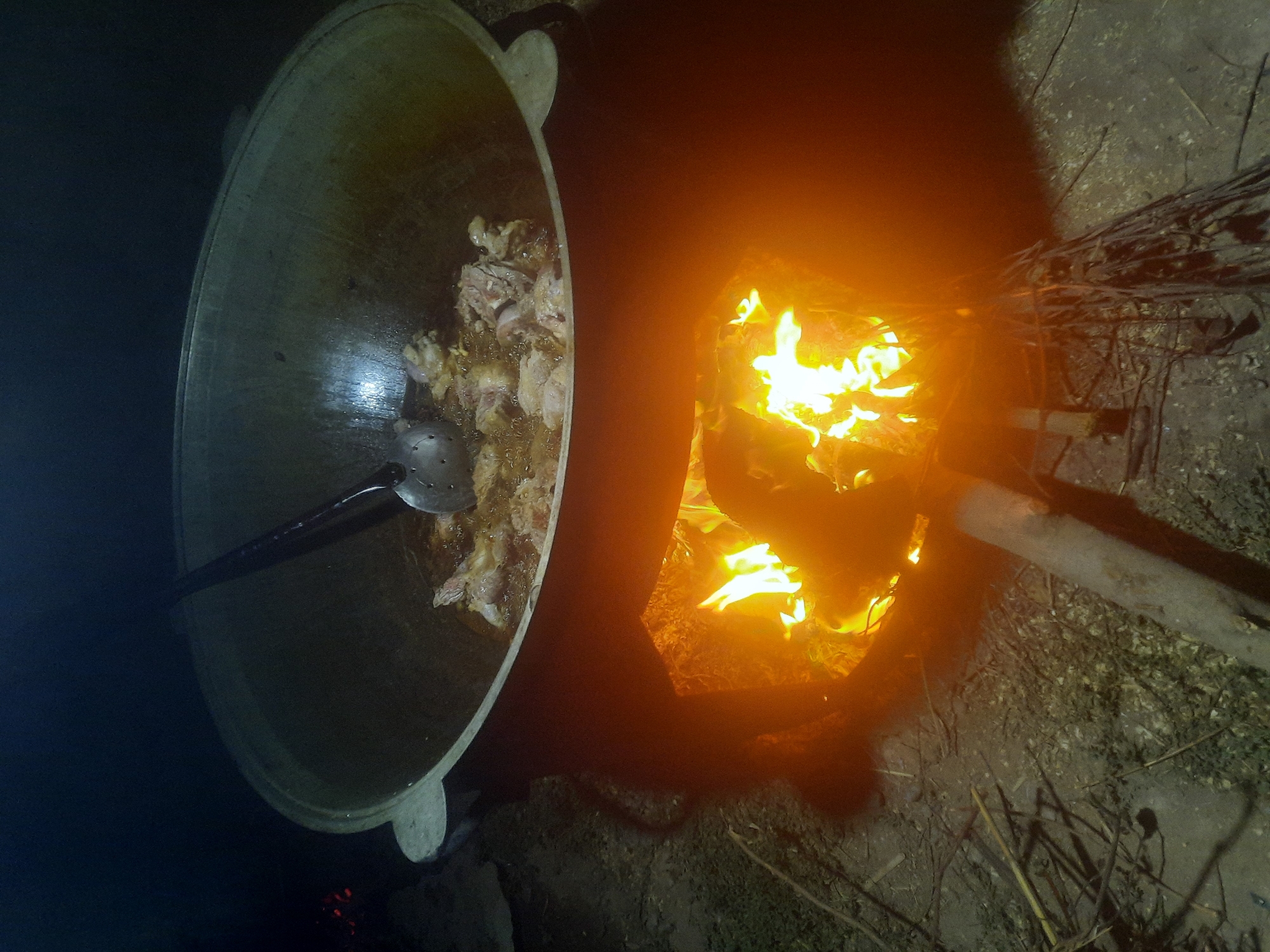 In Восеабад, Tajikistan, an outdoor cooking scene unfolds at night. A large, cast-iron cauldron, likely a kazan, is positioned over a blazing fire built within a makeshift brick or clay oven. The fire casts an intense orange glow upwards, illuminating the contents of the cauldron. Inside the cauldron, pieces of meat, possibly chicken or lamb, are frying in hot oil, with a slotted spoon resting on the edge, ready for use. The surrounding ground appears to be dirt and sparse dry brush, indicating a rural or open-air setting. The darkness of the night contrasts sharply with the bright flames and the cooking activity.