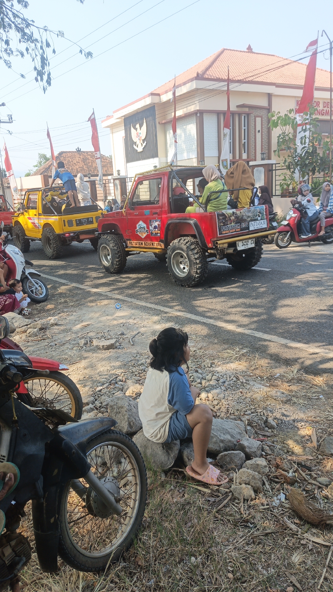 In the city of Kandangmas, Indonesia, on a sunlit day, several red and yellow open-top off-road vehicles are parked alongside a road. The vehicles are decorated with red and white Indonesian flags. A building with a red tiled roof and a large emblem of an eagle stands behind the vehicles. Several people are seen in and around the vehicles, some of whom appear to be spectators. A young girl is sitting on rocks on the side of the road, facing away from the camera, watching the scene. In the foreground, the wheels of a black motorcycle are visible. The environment is dry and dusty, with scattered rocks and vegetation.