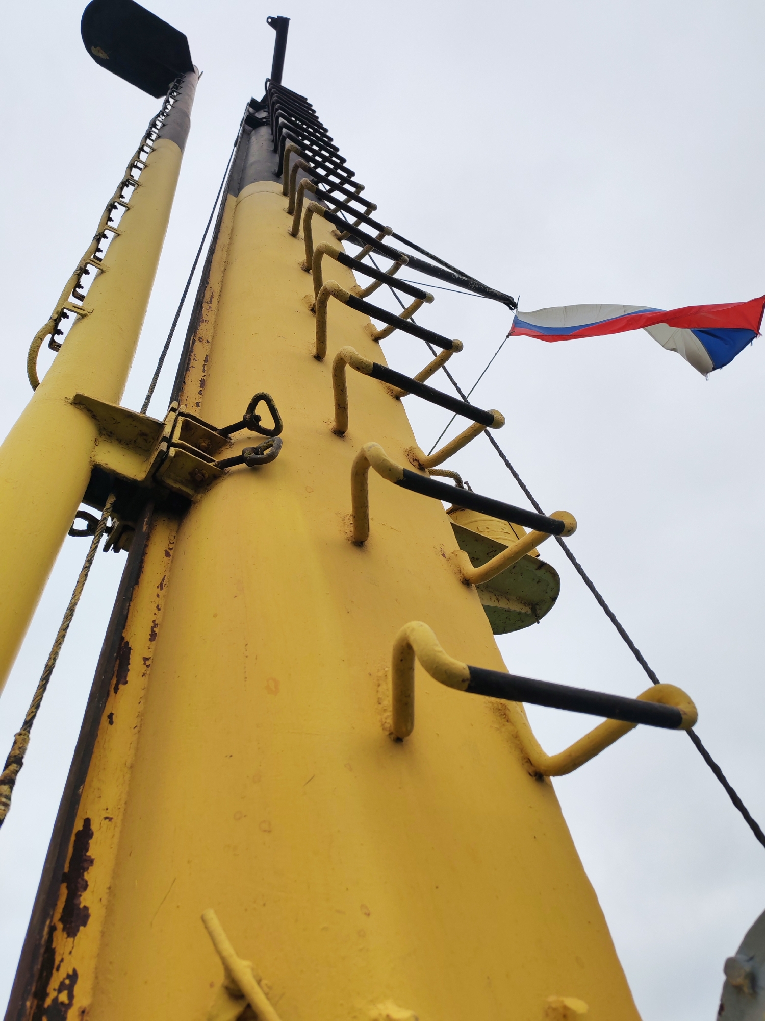 The image depicts a low-angle view of a yellow metal structure with a ladder and a Russian flag at the top. The structure appears to be some kind of mast or tower. The sky is overcast. The image likely originates from Radchenko, Russia, based on the flag's presence.