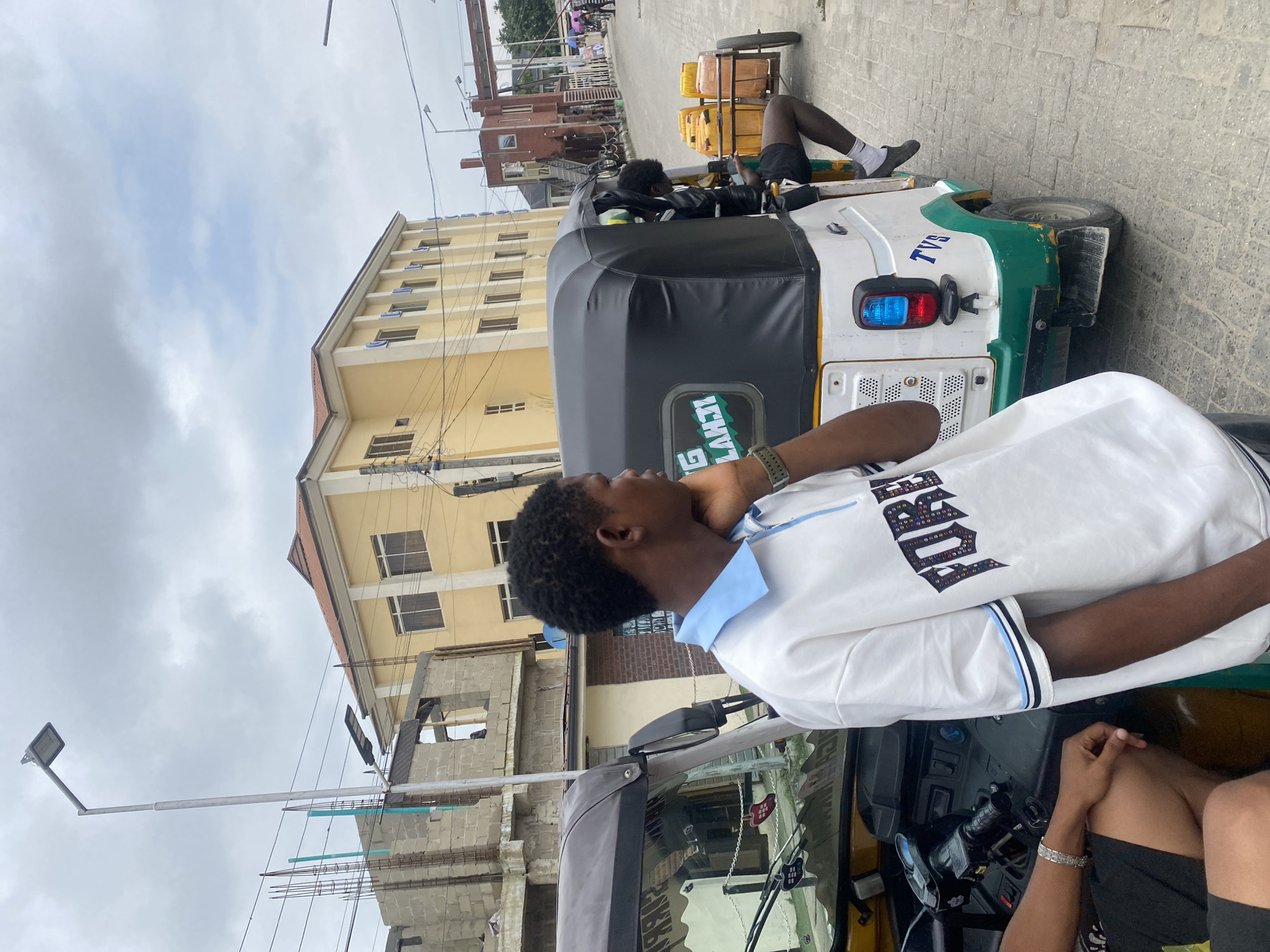 In Unknown, Unknown, a young man stands near a white and green auto-rickshaw. He is wearing a white polo shirt with blue trim and the word "FOREVER" in colorful lettering across the chest. His left hand rests on his chin as he looks to his right, away from the camera. The auto-rickshaw has a black canopy, and another person is visible sitting in the back, legs exposed. The rickshaw is loaded with several yellow containers on a trailer. In the background, a yellow building with multiple windows stands under a cloudy sky. Adjacent to it, a partially constructed building with exposed bricks and rebar is visible. The ground is paved with interlocking bricks. A street lamp stands to the left of the frame.