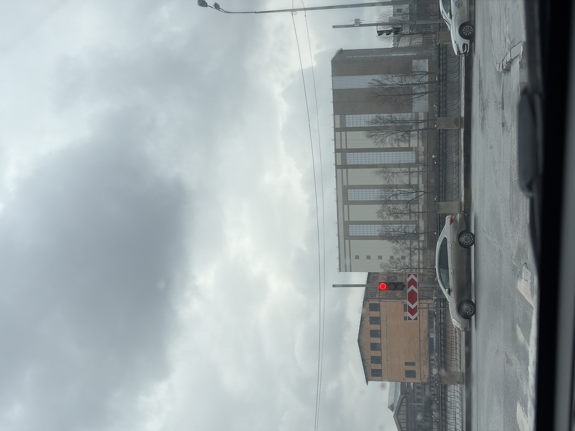 The image depicts a street scene in Saint Petersburg, Russia, taken from inside a vehicle. The sky is overcast, suggesting a cloudy day. A large, gray building with numerous windows dominates the background. In the foreground, there is a traffic light showing a red light, indicating a stop. Next to the traffic light is a building with a light-colored facade and dark window frames. A silver-colored car is stopped on the wet asphalt road. A metal fence runs along the edge of the road. The perspective is from inside a vehicle, as the edge of the window frame is visible.