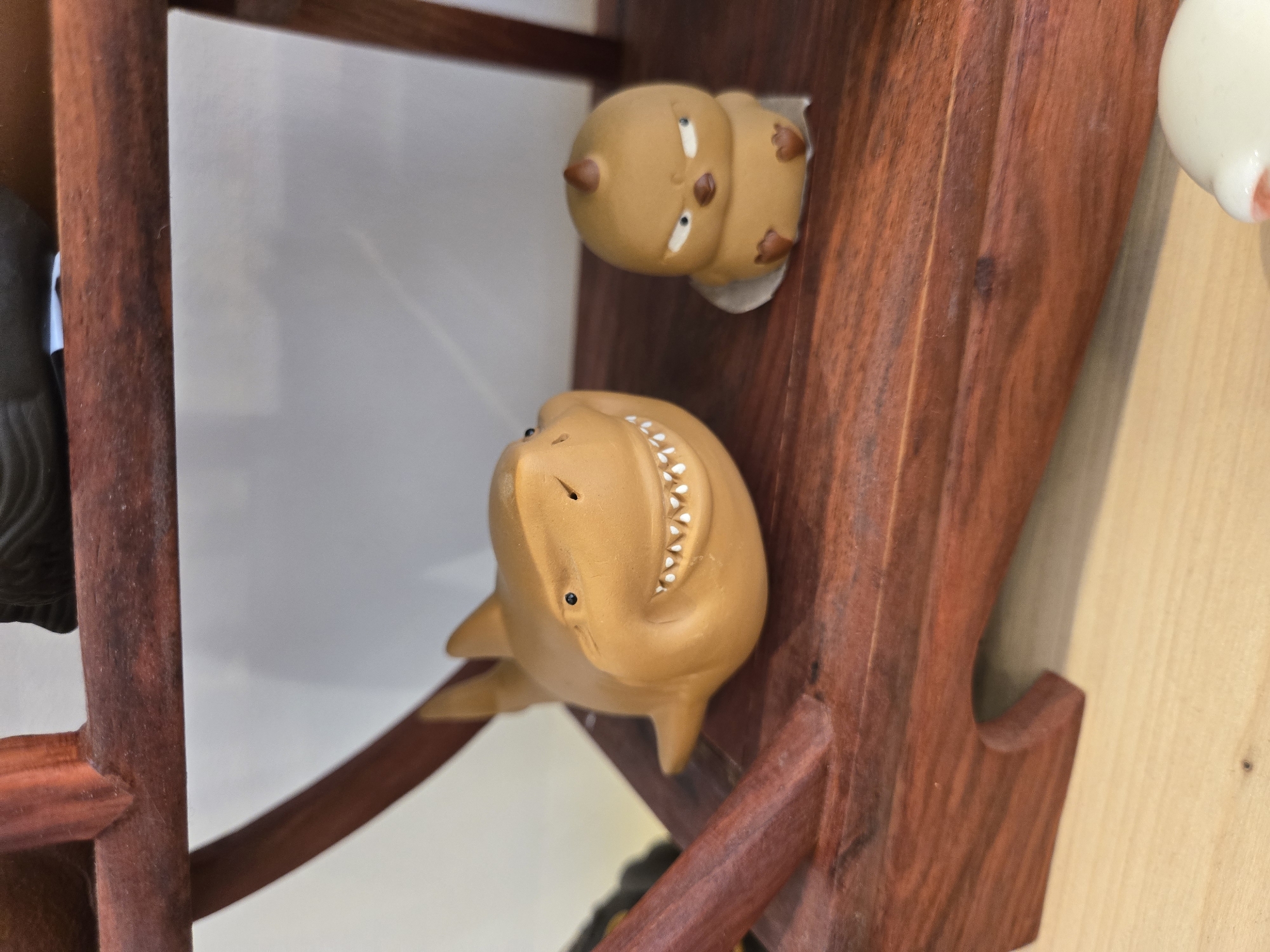 A close-up shot shows two small, brown, stylized figurines placed on a dark wooden shelf. The lower figurine appears to be a shark-like creature with small, white teeth and minimal facial details. Above it, another figurine rests, depicting a squat, rounded figure with cartoonish facial features. The wooden shelf seems to be part of a larger structure, possibly a display case or decorative shelving unit. The background consists of a plain, neutral wall and a light wood surface, suggesting an indoor setting. Based on the user's prompt, the location is assumed to be in Budva, Montenegro, although there are no visual cues within the image to confirm this.