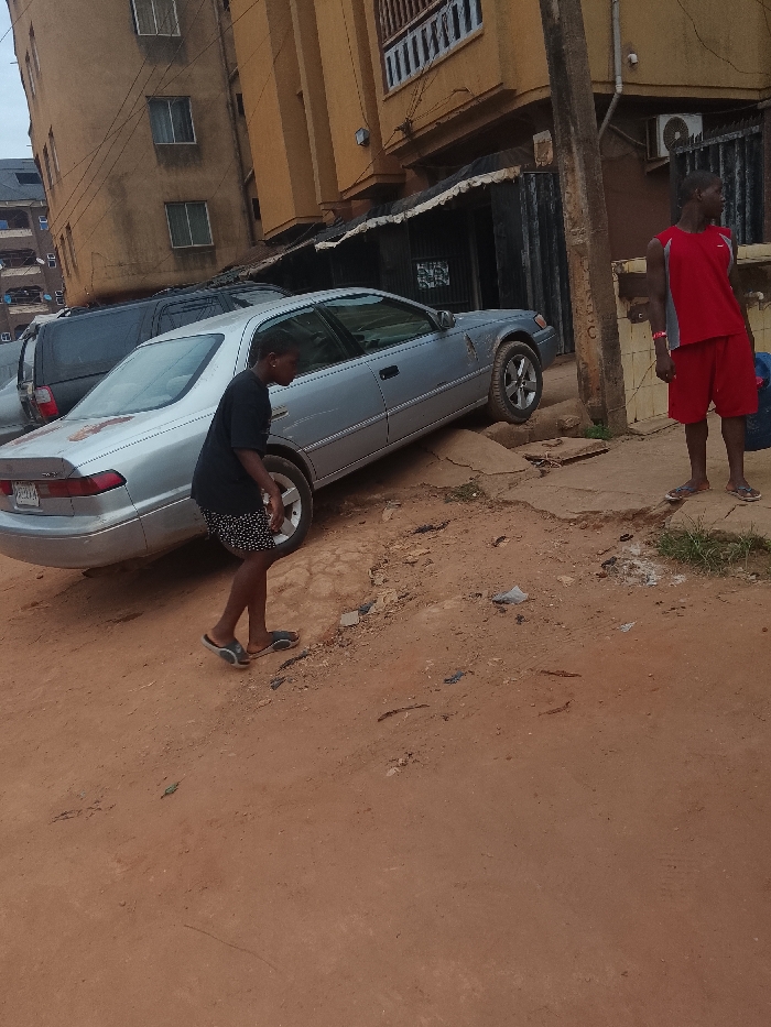 A light-colored sedan sits diagonally on a raised section of a dirt road. Two people are visible. One is walking past the front driver's side of the car wearing a black shirt and patterned shorts. Another person stands near the front passenger's side wearing a red and gray tank top and red shorts. A dark SUV is behind the sedan. Buildings line the background. The road surface appears uneven and broken.