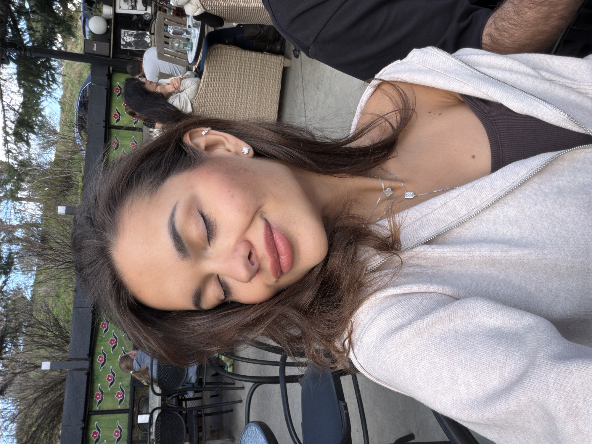 A young woman with closed eyes, light brown hair, and fair skin is in the foreground, wearing a light-colored zippered hoodie and a dark brown top. A silver necklace is visible around her neck. The background features an outdoor cafe setting. Other people are seated at tables in the background, and black metal chairs are in the foreground. Trees, shrubs, and some buildings are also visible in the background. The location appears to be in Tbilisi, Georgia, based on the general architecture, vegetation, and atmosphere reminiscent of the city.