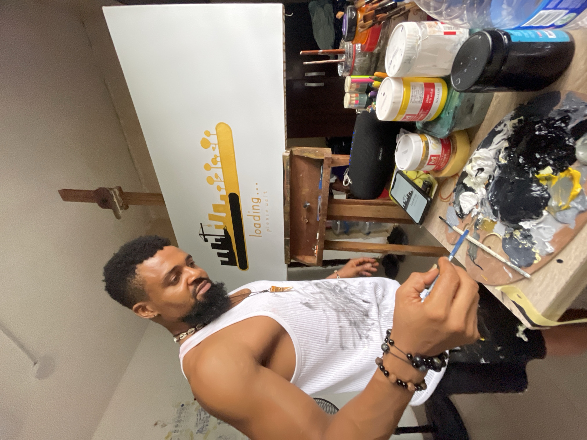 A man is painting in a room. The man is Black, has a beard, and is wearing a white tank top and beaded bracelets. He holds a paintbrush and palette. Several jars of paint are in the background, along with paintbrushes and a speaker. An easel is present with a white canvas that has a yellow graphic and the words "loading... please wait." The room is indoors, and the setting seems to be Ado, Nigeria, though specific location markers are absent. There is no information that confirms this location.