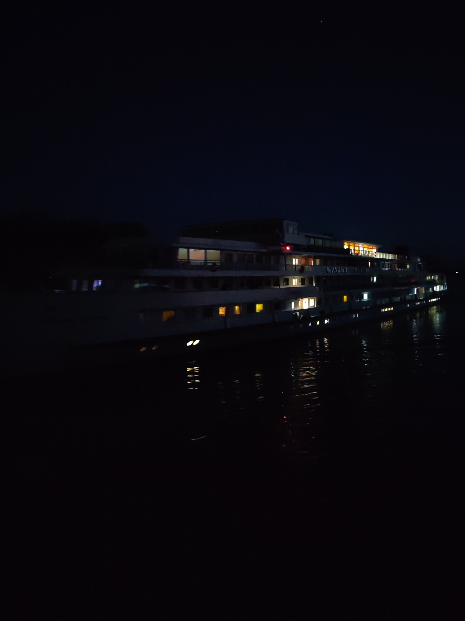 A large, multi-decked riverboat is illuminated by numerous windows and interior lights as it rests on dark, calm water at night. The boat is predominantly white with a darker upper deck structure. The lights create reflections on the water's surface. The sky is a deep, dark blue with a single star visible. The general ambiance is tranquil and still, characteristic of a night scene in a rural or less urbanized setting. The vessel appears to be docked or anchored, as there is no visible movement. The image has a dark, low-light quality due to the time of day.