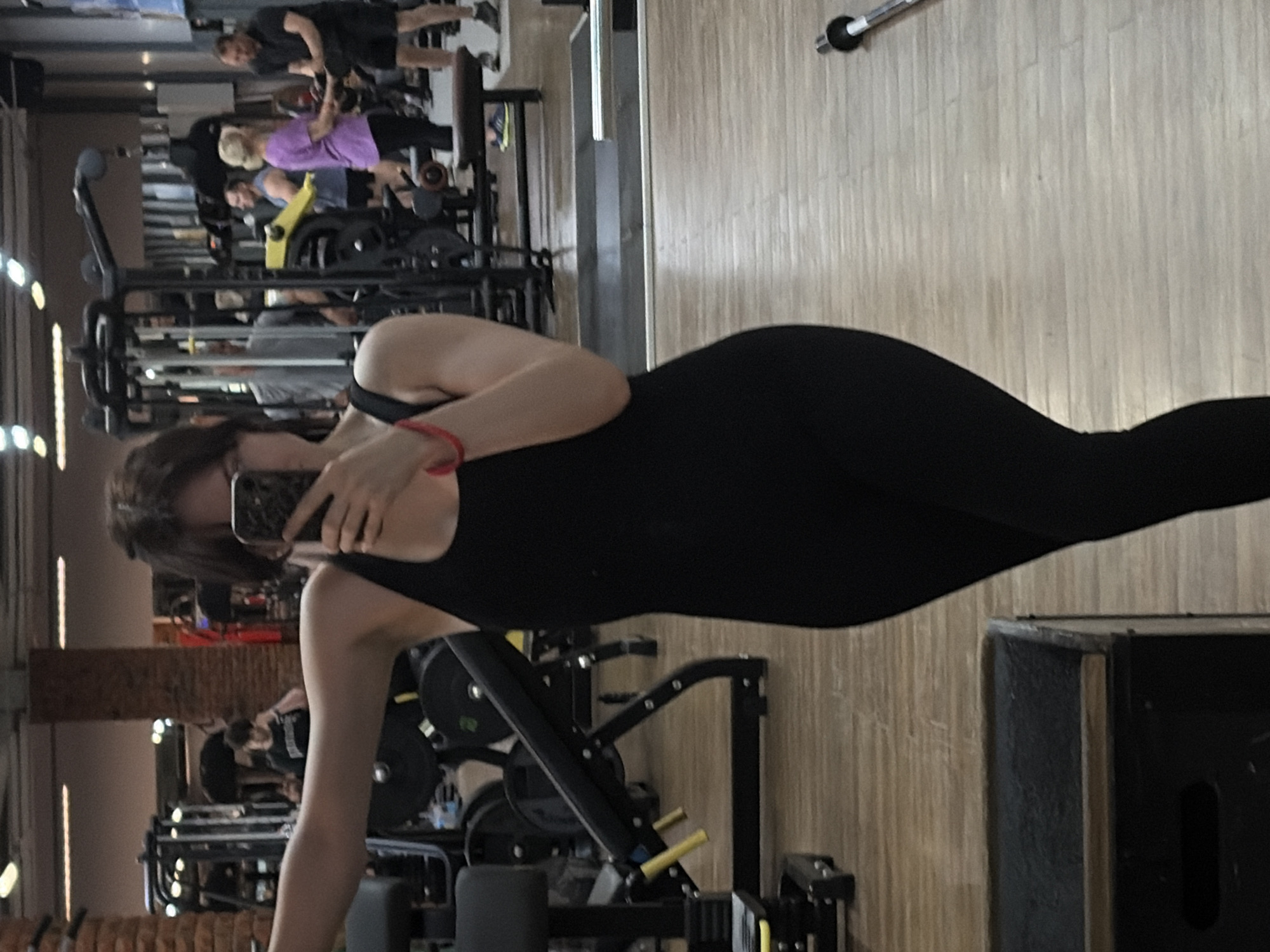 The image is a full-body selfie taken in a gym. A woman with dark hair, wearing a black one-piece workout outfit and a red bracelet on her left wrist, is holding a smartphone and taking a picture of herself in a mirror. She is standing in the foreground, slightly off-center to the left.
The background shows a typical gym environment with various weightlifting equipment, including weight machines, benches, and a barbell on the floor. Several other people are visible in the background, engaged in their workouts or moving around the gym. The flooring is light-colored wood. The gym appears to be indoors and well-lit by overhead lights. The specific location in the city of Chaikovsky, Russia, is not discernible from the image itself.