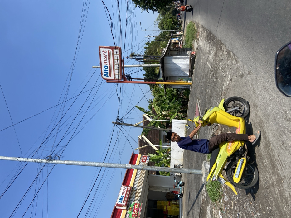 A man wearing a black skullcap is sitting on a yellow scooter parked on a street in Mojosari, Indonesia, at -7.503 latitude and 112.541 longitude. Behind him, a red building with the sign "Alfamart" is visible, and there are a number of electrical wires running overhead. Further back, another Alfamart sign is visible on a pole, and another, smaller building with the sign "ATM" is visible. The man's scooter is facing forward, and he is looking towards the right. The ground around the scooter is covered in gray asphalt. The street is lined with a number of trees and bushes. The sky is blue and clear.