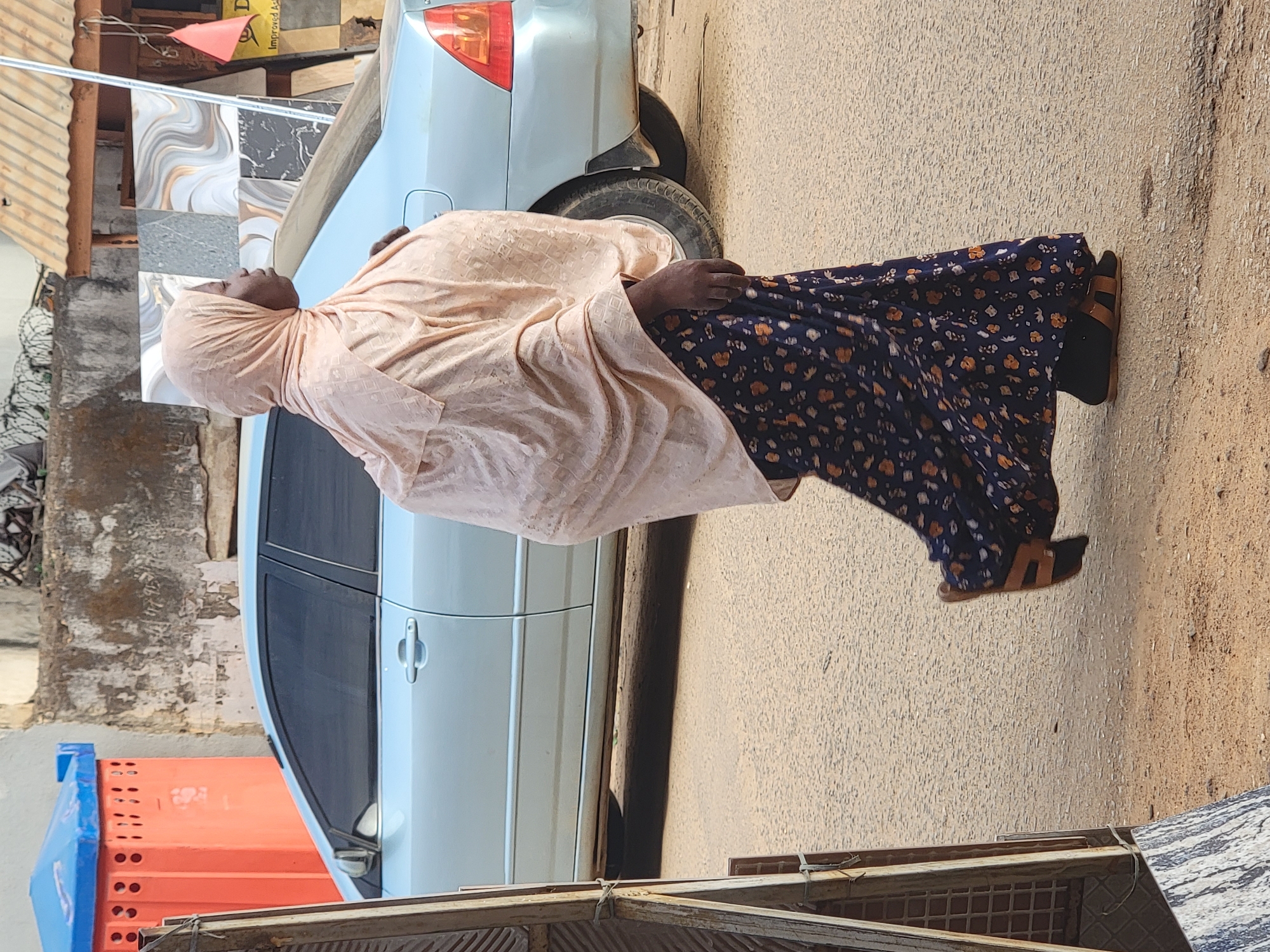 A woman wearing a peach-colored head covering and a long, dark blue floral dress stands next to a light blue car on a paved street. She is wearing tan sandals over black socks. The car is parked along the side of a building that appears to be under construction or renovation, as evidenced by stacked orange crates and a corrugated metal roof. Tiles of varying patterns are stacked near the car. The location appears to be Maiburiji, Nigeria, indicated by the street's sandy color and surrounding architecture.