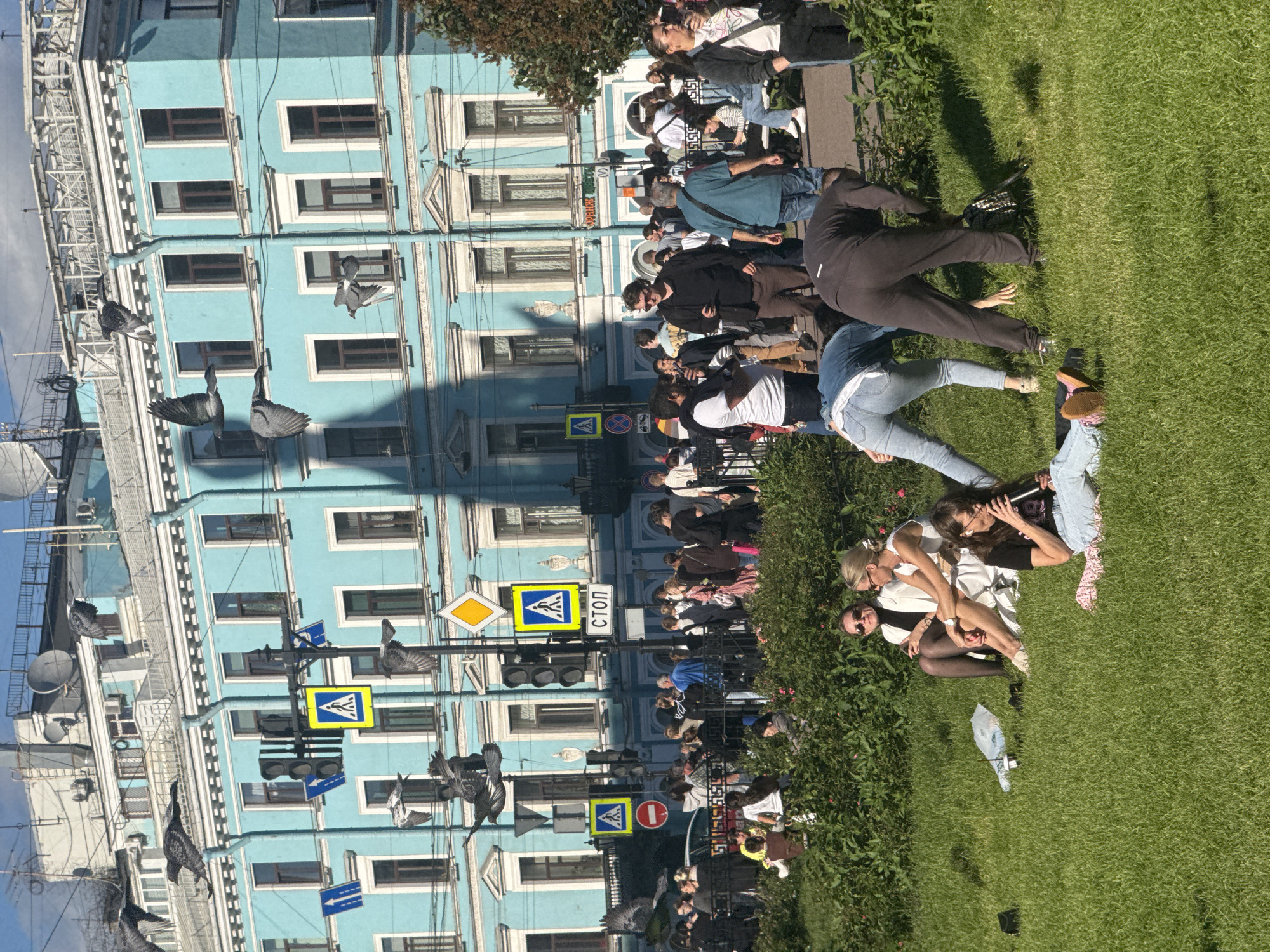 A group of people are gathered on a grassy area in front of a blue building in Saint Petersburg, Russia. The building has multiple windows and architectural details typical of the city. Several pigeons are visible, some in flight and others perched on wires or structures. The scene appears to be outdoors on a sunny day, with shadows cast on the grass. Some individuals are sitting or lying on the grass, interacting with each other, while others are standing and observing. The overall atmosphere seems relaxed and informal. Traffic lights and street signs are present, including a pedestrian crossing sign and a diamond-shaped yellow sign with a black border. There is also a red circular sign with a white horizontal bar, likely a "stop" sign or prohibition marker. The word "СТОП" (STOP) is visible on one of the signs. The image is oriented sideways.