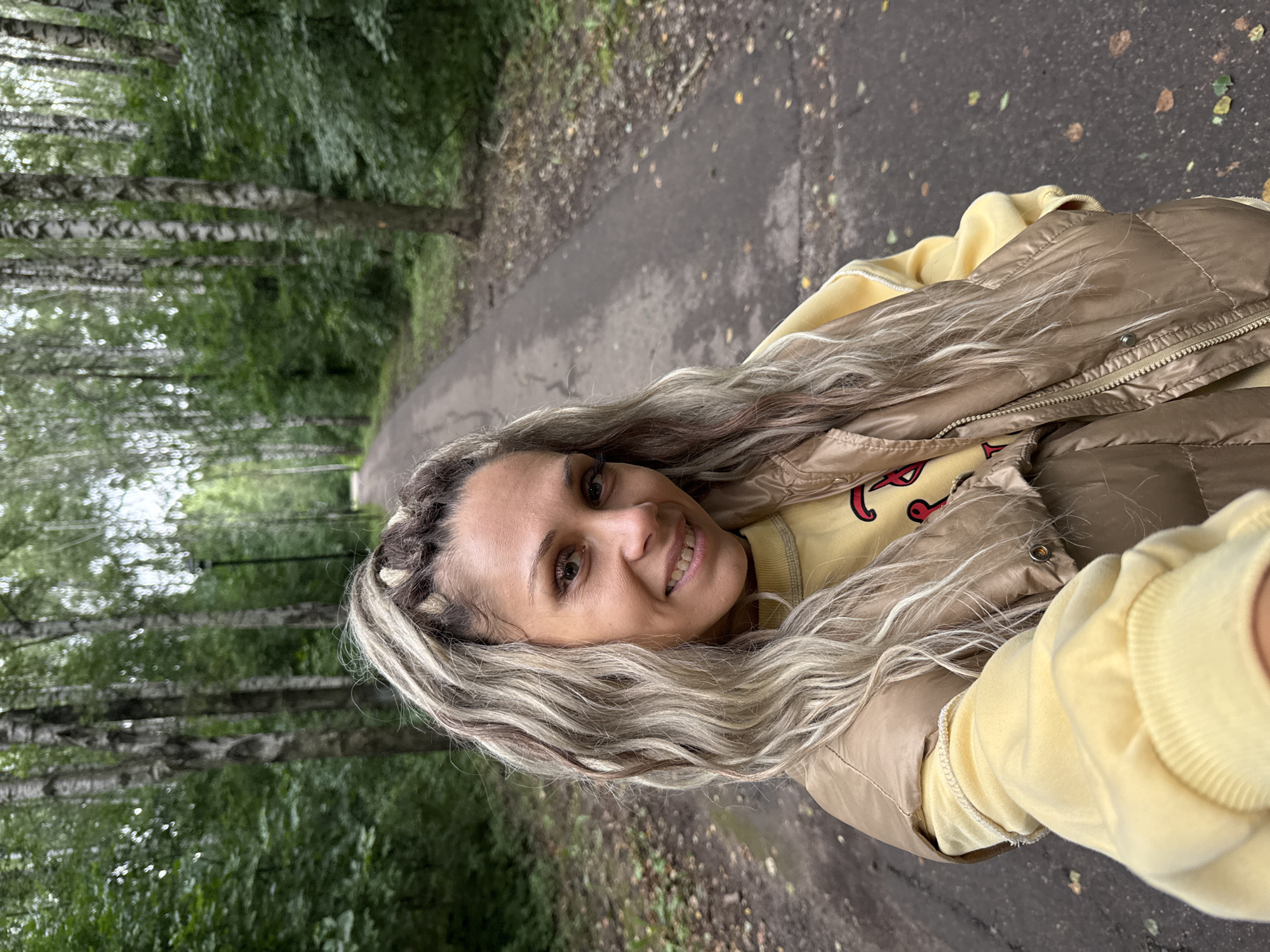 In Rusanovo, Russia, a woman with blonde and brown braided hair styled in a high ponytail is captured in a selfie outdoors on a paved path. She is smiling and looking directly at the camera. The woman is wearing a yellow sweatshirt under a metallic gold puffer vest. The path is surrounded by a dense forest with tall birch trees and lush green foliage. The ground on either side of the path is covered with fallen leaves. The lighting suggests it is daytime, possibly overcast.