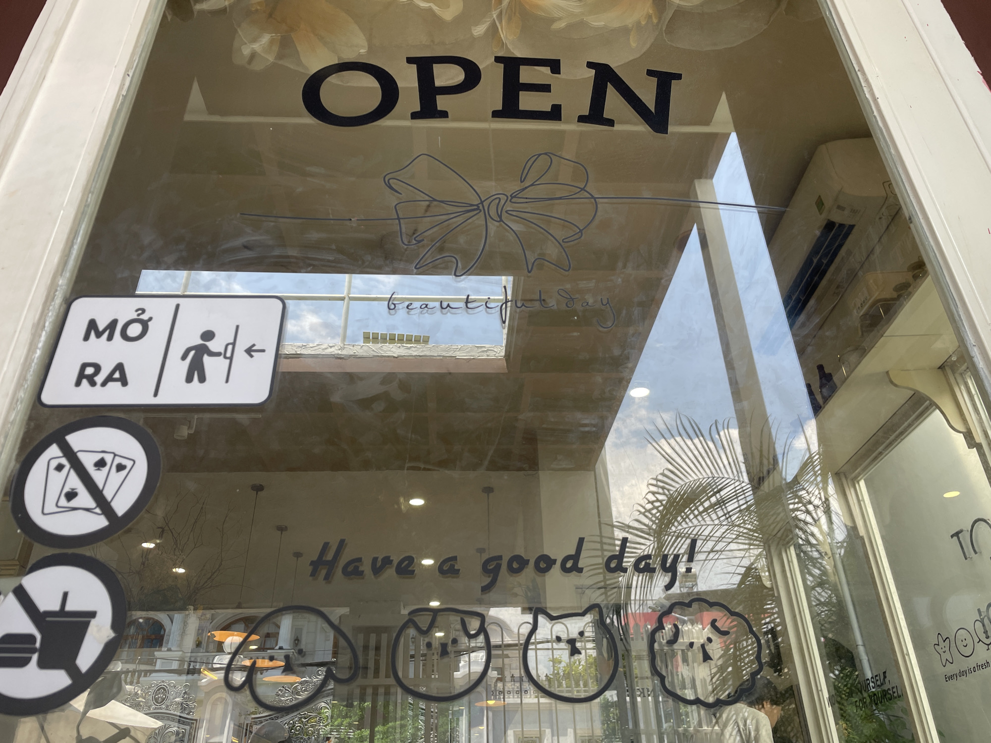 The image shows the front window of a establishment, likely a cafe or shop, in Nha Trang Ward, Vietnam. The word "OPEN" is prominently displayed in large black letters across the top of the window. Below that, a sketched bow is visible, followed by the handwritten words "beautiful day".
On the left side of the window, there is a sign with "MỞ RA" which translates to "OPEN" in Vietnamese, next to a pictogram of a person opening a door. Below this are two circular prohibition signs: one with playing cards crossed out, indicating no gambling, and another with a burger and a drink crossed out, suggesting no outside food or drinks.
Towards the bottom of the window, the phrase "Have a good day!" is written in a script font. There are also several cartoonish animal face outlines drawn on the glass, including what appears to be a dog, a cat, and another animal.
Reflections on the glass reveal the sky, palm trees, and parts of buildings across the street. An air conditioning unit is visible on the ceiling inside the establishment. The overall impression is a welcoming and perhaps whimsical storefront.