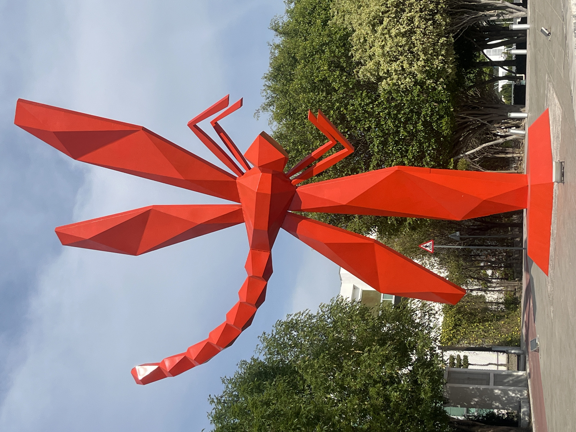 A large, vibrant red sculpture of a dragonfly dominates the scene, positioned outdoors against a backdrop of lush green foliage and a partly cloudy sky. The sculpture is highly geometric, constructed from angular, faceted planes that give it a modern, abstract appearance. It features a distinct head, a multi-segmented body and tail, and prominent, broad wings that also exhibit the same angular design.
The setting appears to be a public outdoor space, possibly a park, plaza, or a roadside area in an urban or suburban environment. Dense green trees with varied foliage, some lighter green or yellowish, form the background, partially obscuring a building. A paved surface, possibly a sidewalk or pathway, is visible in the foreground and extends along the base of the sculpture.
No human activity or people are present; the scene is static, focusing solely on the sculpture and its immediate environment. The bright illumination suggests it is daytime. A notable detail observed through the trees is a triangular road sign with a red border, depicting a black pedestrian symbol on a white background, indicating a pedestrian crossing or children playing nearby. The original image is oriented vertically, making the dragonfly appear upright, though its natural orientation would be horizontal.