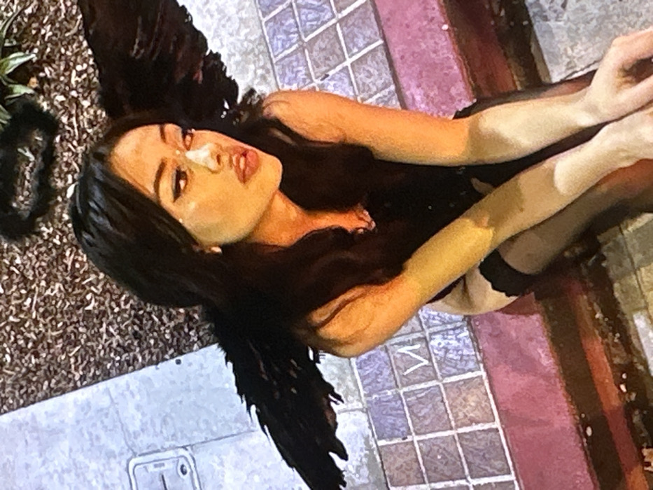 A young female, with long dark hair and dark winged makeup, is depicted in a seated or crouched position. She is wearing a black corset-style top or lingerie, black thigh-high stockings, and large black feathered wings attached to her back. Small white decorative elements are visible below her right eye and on the bridge of her nose. Her lips are slightly parted, and she gazes towards her right (viewer's left). Her hands are clasped together in front of her. She is situated on a low, red-painted curb or wall. To her left is gray concrete pavement with a rectangular utility cover. To her right on the ground, there is patterned tile flooring, one tile distinctly featuring a Cyrillic letter "И". Behind her, a dark, textured ground surface with green foliage is visible, along with a fuzzy black halo-like object. The scene takes place in the city of Novosergievka, Russia.