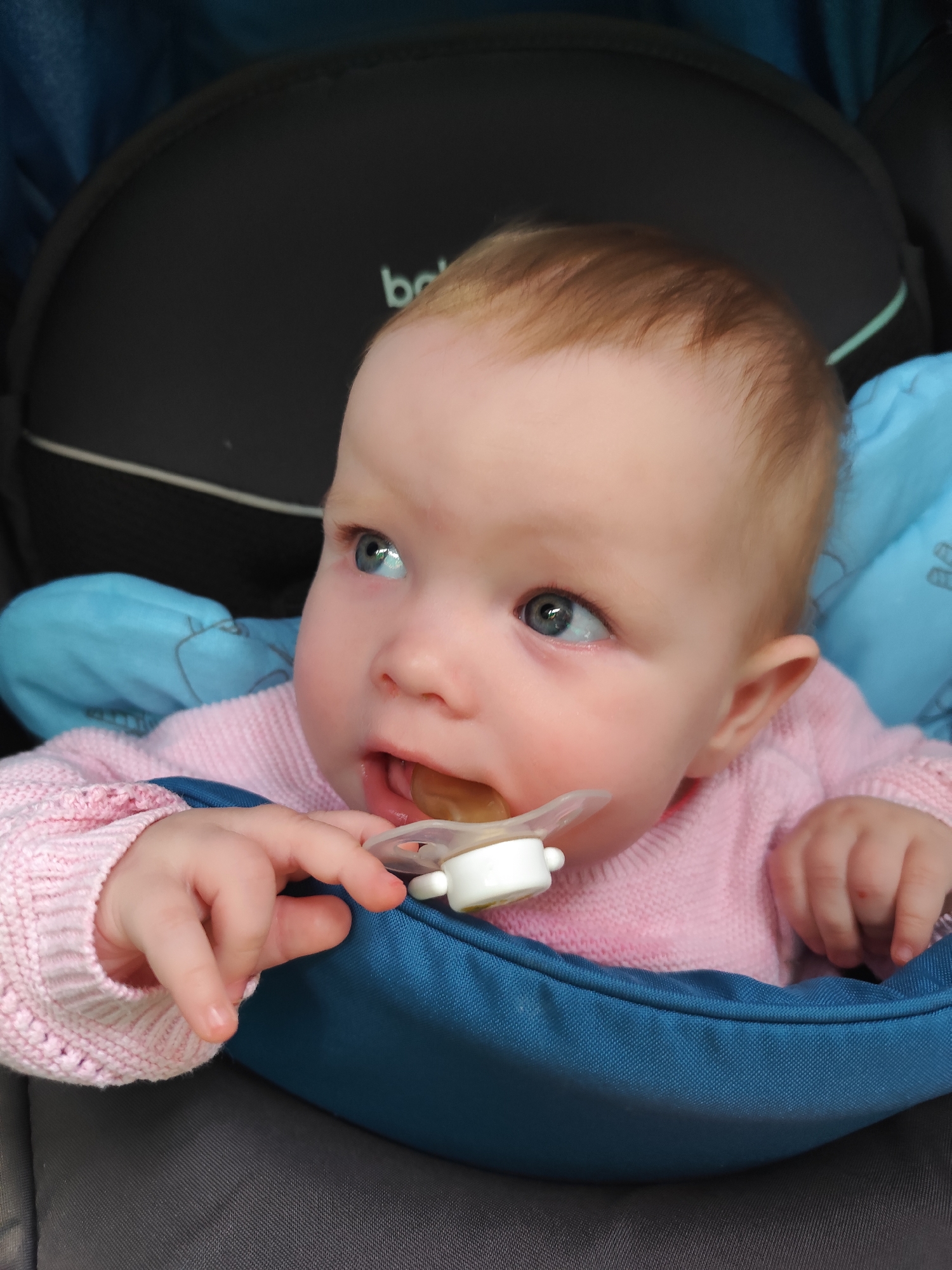 A baby with fair skin and light reddish-blonde hair is seated in a stroller. The baby is wearing a pink knitted sweater and has a pacifier in their mouth, held by one hand. The stroller has a black padded back, a teal-colored cushion, and a dark gray bottom. The background is slightly out of focus. The photo does not depict the city of Chaikovsky, Russia.