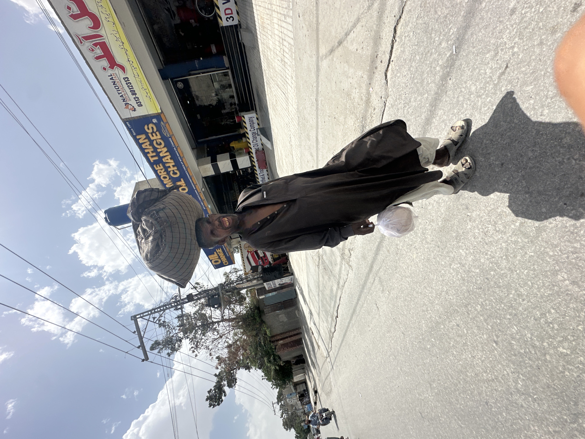 A man stands on the sidewalk in Quetta City Tehsil, Pakistan. He wears a dark brown, long-sleeved traditional garment and light-colored sandals. He is carrying a large woven basket-like object on his head and a white plastic bag in his hand. A sign for "NATIONAL OILS PAKISTAN" is visible behind him, advertising oil changes and listing a phone number. The street is made of concrete, and power lines are visible against a backdrop of a partly cloudy sky. Other buildings and foliage are present in the background, adding context to the urban scene.