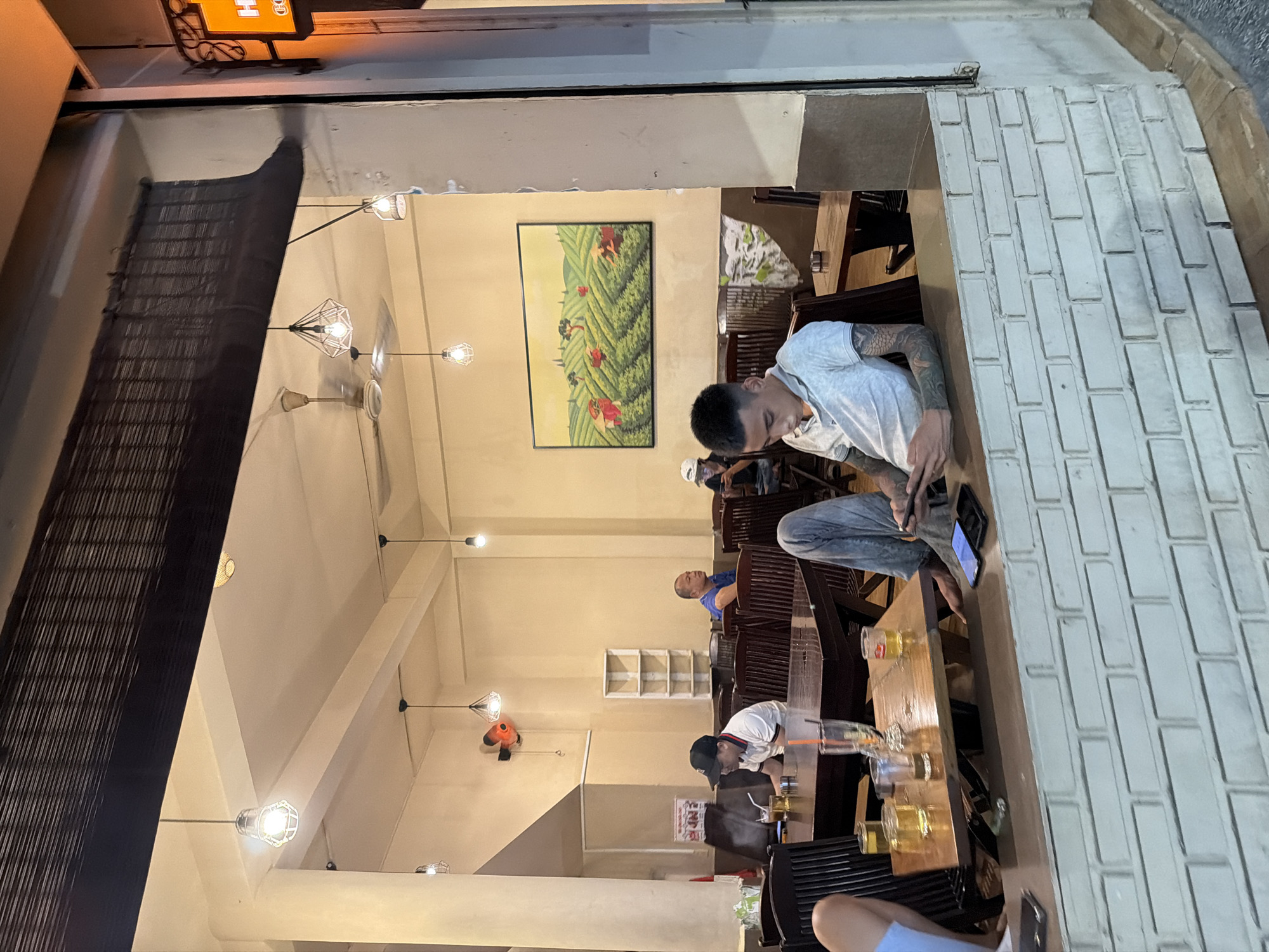 The image shows the interior of a cafe in Nha Trang Ward, Vietnam, viewed from outside through a large window. Inside, several people are seated at wooden tables. A young man in the foreground is looking down at his phone. He is wearing a light-colored t-shirt and has tattoos on his arms. Other patrons are visible in the background, including a man wearing a blue shirt and a man wearing a black baseball cap. The cafe has a cream-colored ceiling with multiple pendant lights and a ceiling fan. A large framed painting of a landscape with figures harvesting crops hangs on the wall. The foreground is dominated by a white brick counter.