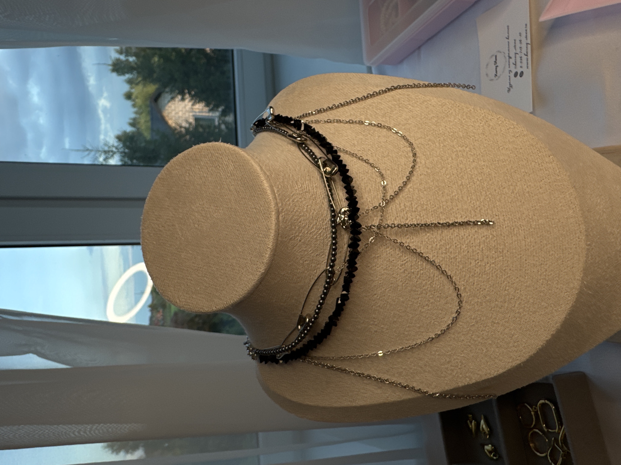This media file features a mannequin bust adorned with layered necklaces, including one with black beads and safety pin embellishments and another made of delicate silver chains. The bust is positioned in front of a window offering a view of trees and a cloudy sky, suggesting it might be outdoors or near a window with natural light. The background also shows hints of white curtains and a box containing jewelry, possibly indicating a retail or display setting. The time of day appears to be overcast, and there are no people visible in the frame. The text visible on a card in the background is in Russian and includes a website and phone number.