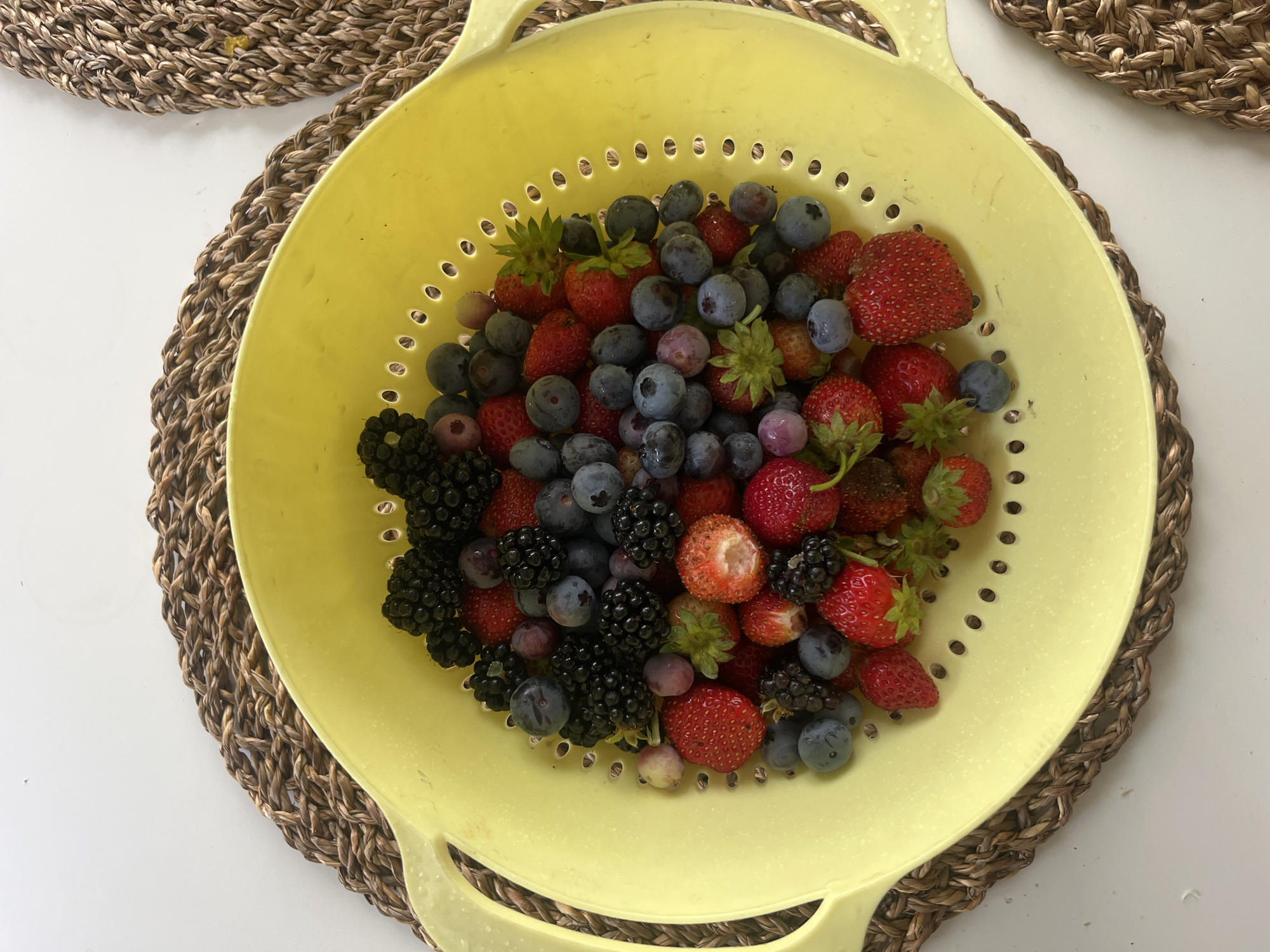 A bright yellow colander filled with fresh mixed berries, including strawberries, blueberries, and blackberries, is placed on a woven placemat. The placemat is on a white surface, possibly a table. The colander has several drainage holes. Some of the berries are whole, while others appear to have been bitten into, revealing their juicy interiors. The overall impression is one of a healthy and appetizing selection of summer fruits. There is no indication of a specific location within the city of Столбище, Russia.