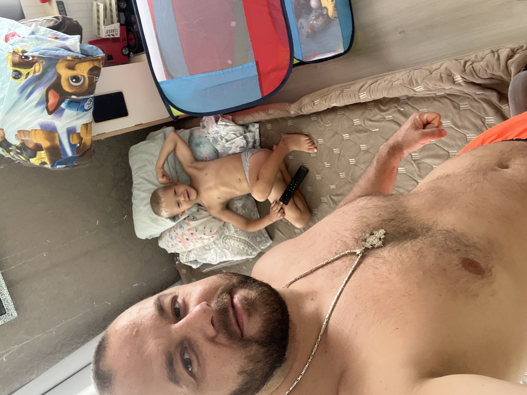 The image shows a man and a young child lying on a bed in what appears to be a bedroom. The man is in the foreground, taking a selfie. He has a beard and is wearing a silver necklace. The child is lying on a pillow behind him, also on the bed. The child is wearing a diaper and appears to be holding a remote control.
In the background, there are various objects visible, including a playpen with colorful panels, a table with a tablet, and what looks like children's toys, possibly including a red toy truck. The bed has a patterned comforter. The room is indoors and the lighting suggests it is daytime. The overall scene conveys a casual, domestic setting.
The image appears to be taken in Sochi, Russia. There is no visible text in the image. The emotions conveyed are relaxed and possibly playful, given the selfie pose and the presence of the child.