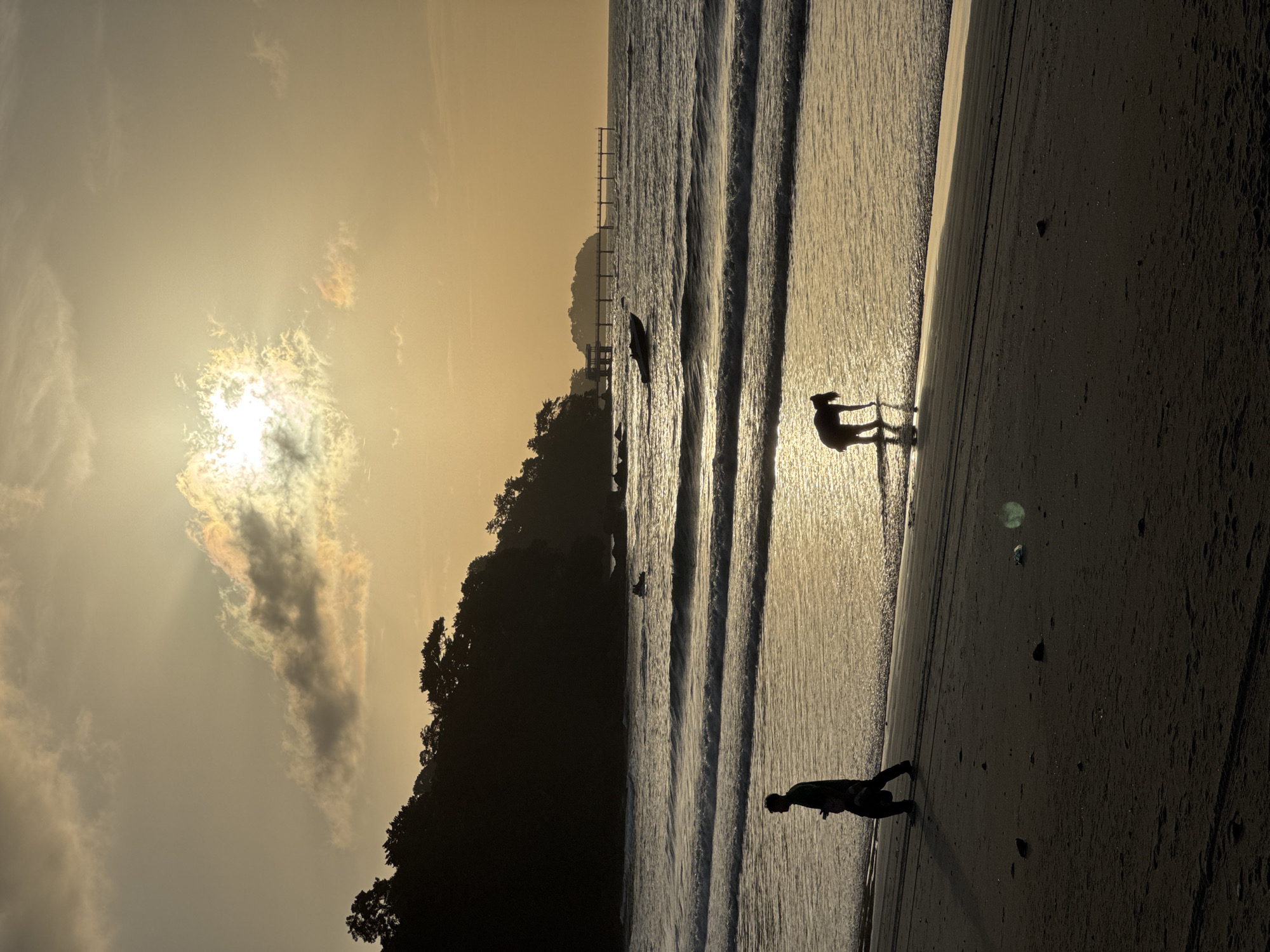 A beach scene in Patong, Thailand, at sunset features two silhouetted figures on the sand and in the shallow water. The sun, partially obscured by clouds with visible crepuscular rays, casts a golden light across the ocean and wet sand. In the foreground, a person walks along the beach, carrying an object in their right hand. Further down the shore, another figure is bent over, appearing to interact with the water. The ocean displays gentle waves breaking near the shore, with a small boat visible in the middle ground. A distant pier extends into the sea from a tree-covered headland on the left. The sand in the foreground is dark and shows tire tracks, indicating it is a public beach.