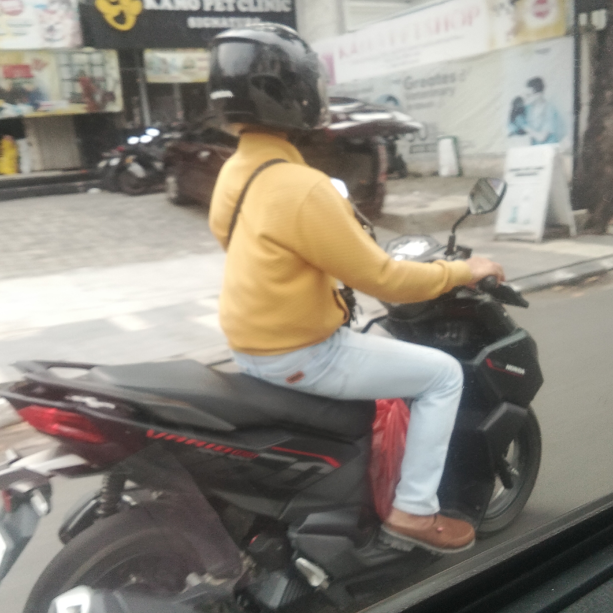 A person wearing a yellow long-sleeved shirt, light blue jeans, and a black helmet rides a black motorcycle. The person is seen from the side, with their right hand on the handlebar. A red bag is visible behind their legs. The motorcycle has a black body with red and white accents. In the background, there are buildings with signs, including one that reads "KANO PET CLINIC" and another that appears to be an advertisement. The scene appears to be on a street in the city of Bogor, Indonesia, with a sidewalk and road visible. The overall image is slightly out of focus.