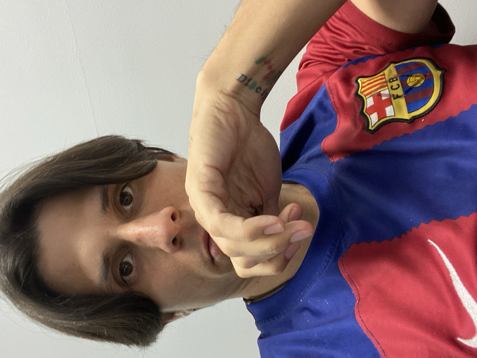 A person in a red and blue FC Barcelona jersey looks at the camera, with a tattoo on their left wrist. Their brown hair is neatly parted, and their hand is positioned near their mouth. The image is a close-up shot taken from below, with a plain white wall in the background. The FCB logo is visible on the jersey's upper right side. A white Nike logo is visible on the bottom right.