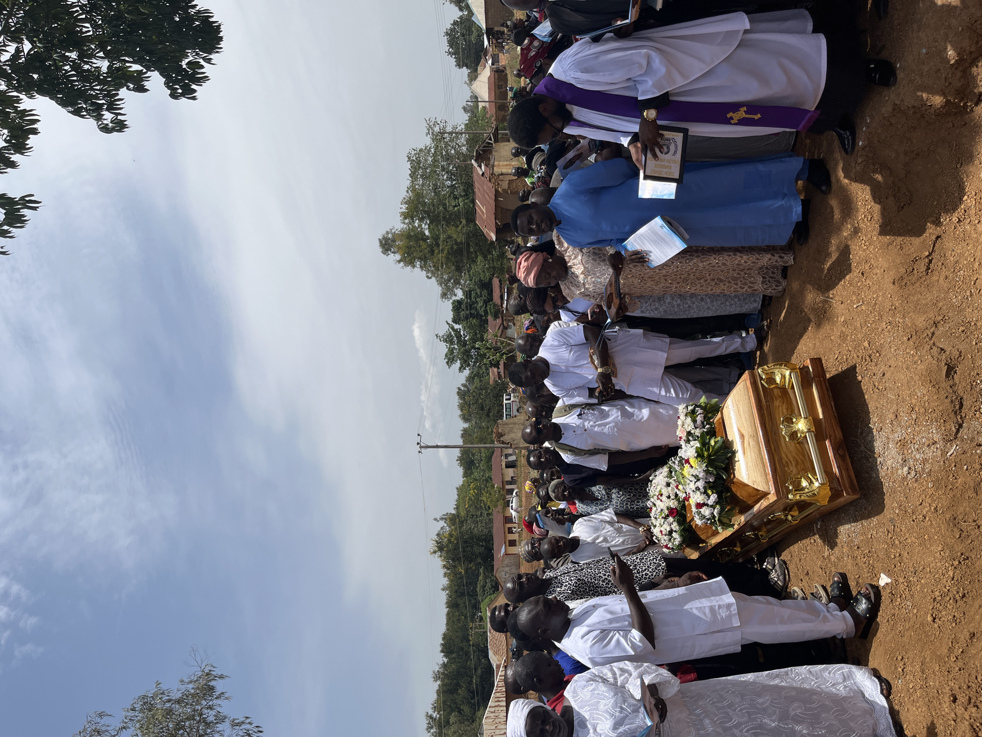 The image shows a funeral in Dogon Dinia, Nigeria. A gold-colored coffin is on the ground, surrounded by people. Many are dressed in white. A clergyman in a white robe with a purple stole stands near the head of the coffin, holding a framed object. Others hold books or papers. The background includes trees, buildings with rusty roofs, and a clear sky. The ground is reddish-brown. People appear to be gathered to mourn and pay their respects.
