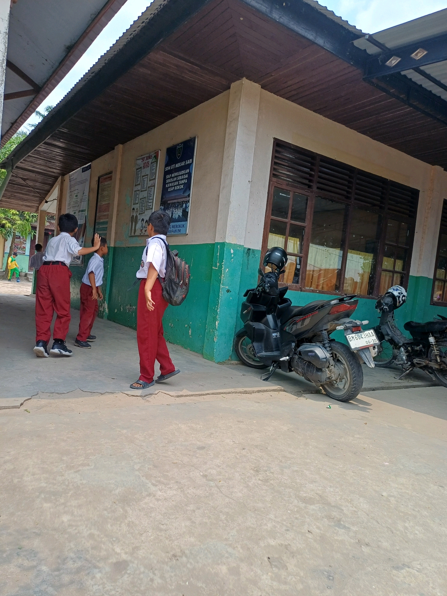 The image depicts a scene outside a school building in Dumai City, Indonesia. The building has a light-colored facade with green painted accents and wooden window frames. A covered walkway extends from the building, supported by pillars, with a corrugated metal roof.
In the foreground, three young boys in red pants and white shirts are visible. One boy, facing away from the camera and wearing a backpack, stands on the right. Another boy, in the center, is facing towards the other two, with his left hand raised as if interacting with the boy on the far left. The boy on the far left is also wearing red pants and a white shirt, and his right hand is raised and touches the back of the boy in the center.
Two motorcycles are parked near the building. The one on the left is black and has a license plate that reads "BM 6043 HAA 06-30". A black helmet is placed on its handlebars. To the right of this motorcycle is another, partially visible, dark-colored motorcycle with a helmet on its handlebars.
Posters are affixed to the wall of the school building, including one with text and graphics related to health or school policies, and another featuring images of children and text. In the background, to the left, a person in a yellow shirt and green pants is visible near a grassy area, and another figure is partially obscured. The ground is paved with concrete. The sky is partly cloudy.