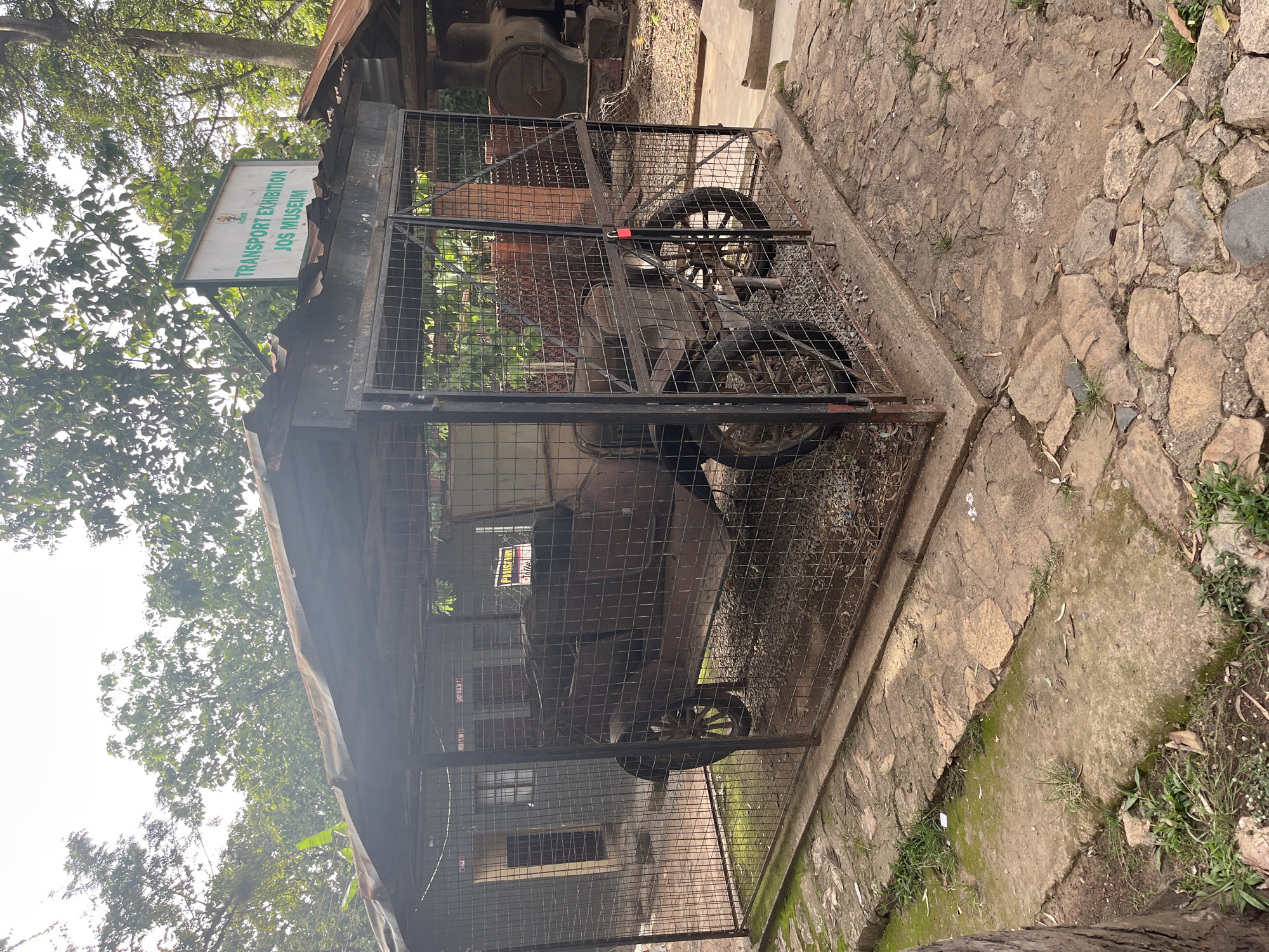 The image depicts an outdoor exhibit at the Jos Museum in Jos, Nigeria. A vintage automobile, likely from the early 20th century, is displayed inside a wire cage. The car is rusted and appears to be in a state of disrepair, with its wooden-spoked wheels visible. Above the cage, a sign reads "TRANSPORT EXHIBITION JOS MUSEUM." The surrounding environment consists of overgrown vegetation, paved pathways, and other outdoor structures, including what appears to be a large steam engine in the background. The scene is bathed in natural light, suggesting it was taken during the day. No people are visible in the image.