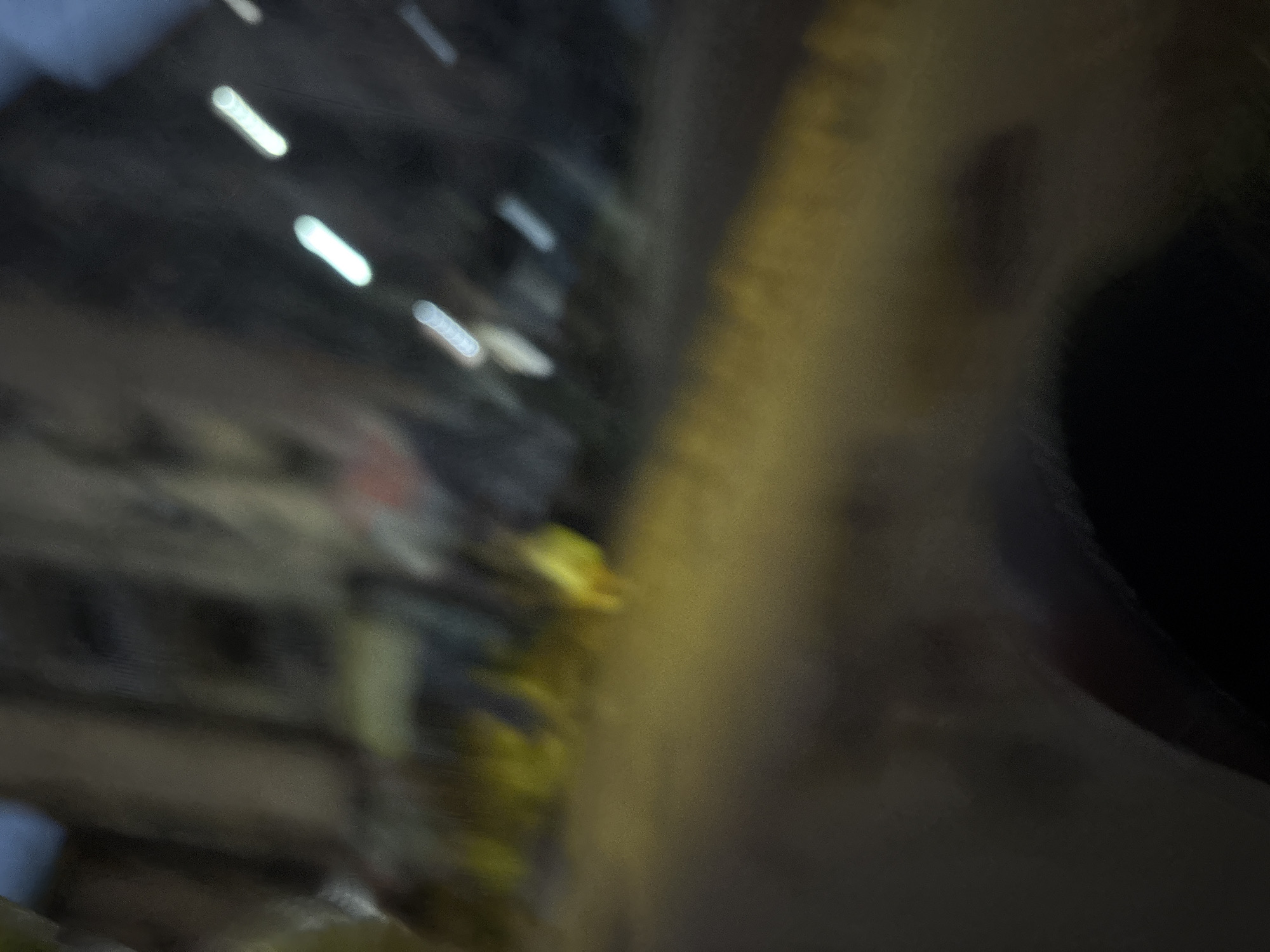 The image, taken at night in Onitsha, Nigeria, shows a blurred street scene with visible streaks of light, suggesting movement, possibly from vehicles. The foreground is dark and indistinct, with a curved, black shape that could be part of a car interior. The middle ground displays a yellowish-brown surface, likely a road or pavement, with some indistinct patches of yellow, possibly indicating streetlights or reflective markings. In the background, the blurred shapes of buildings are visible, with several elongated white lights streaking across them, further emphasizing the nocturnal and mobile nature of the scene. A small, indistinct yellow form is visible in the middle ground, potentially a person or an object illuminated by a light source. The overall impression is one of a dynamic urban environment at night, captured with a sense of motion blur.
