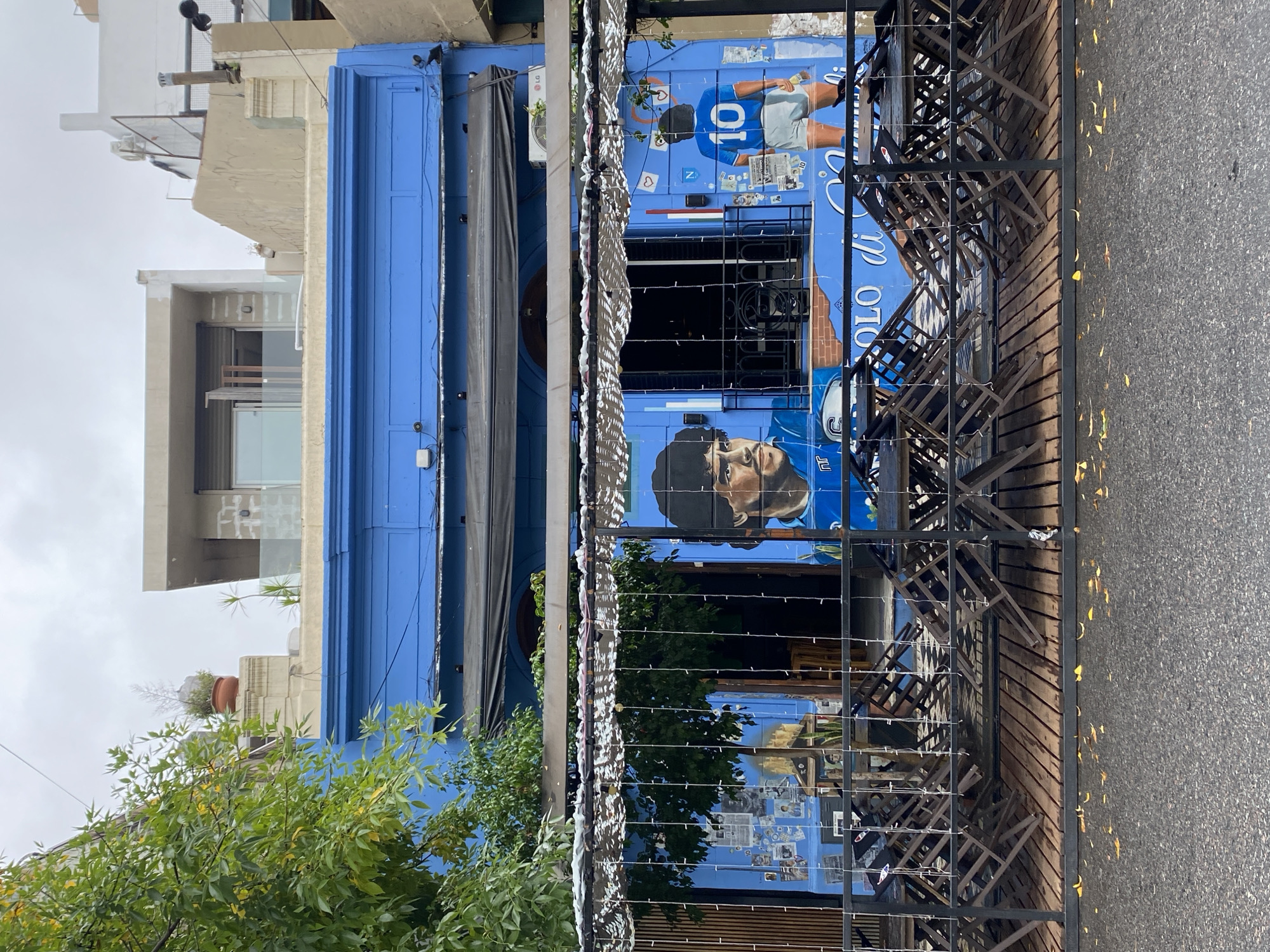 The image captures a vibrant street scene in Buenos Aires, Argentina, featuring a building facade painted in a bright blue color. The facade is adorned with mural-style depictions of Diego Maradona, a famous Argentinian soccer player. A black metal fence partially obscures the view of the building. The street in front of the building is visible, and there are some leaves scattered on the pavement. The sky is overcast. There are stacked chairs within the fence. Vegetation is visible on the left side of the image. Other buildings are visible in the background.