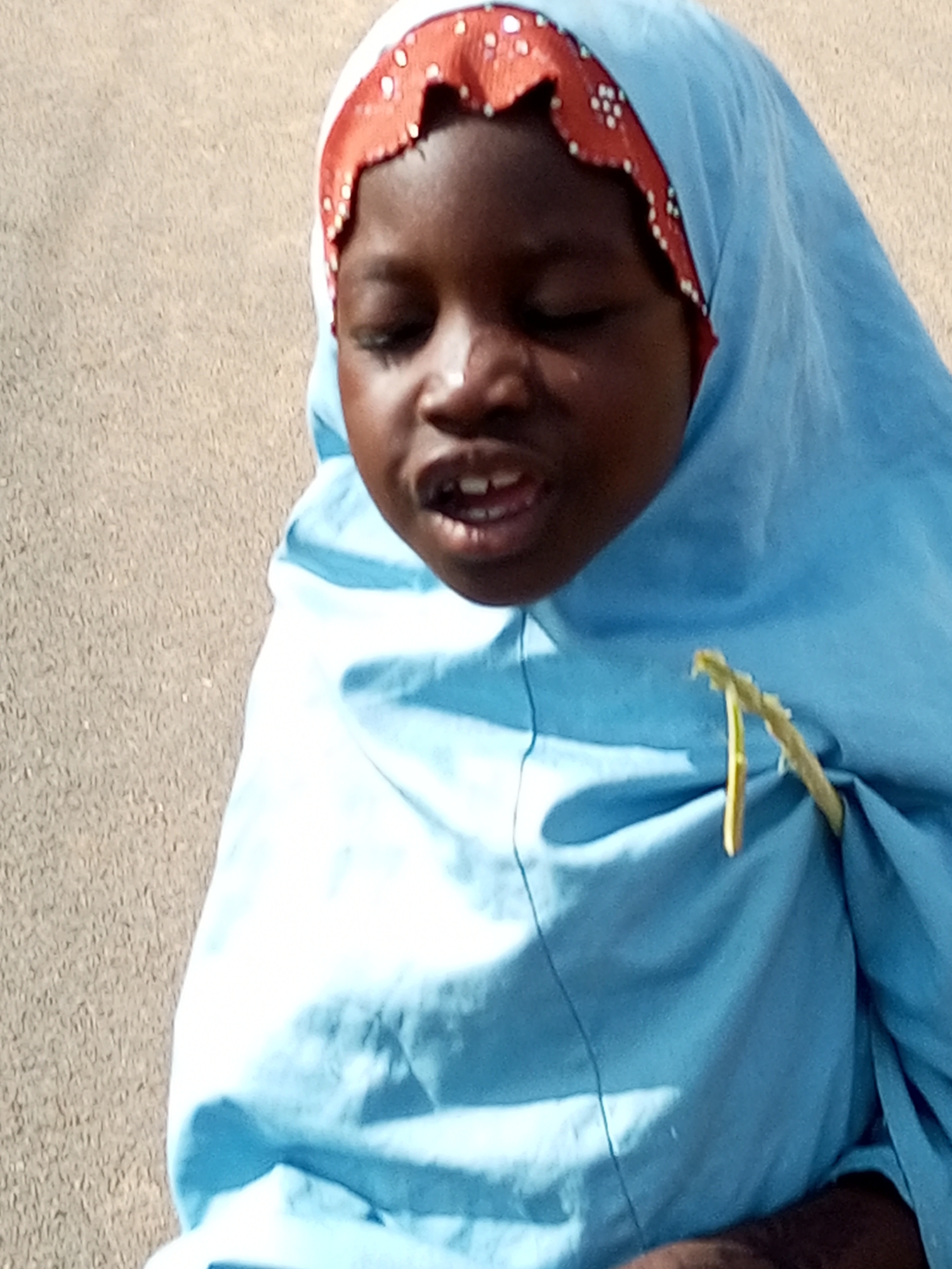 In Gombe, Nigeria, a close-up, eye-level, outdoor shot focuses on a young Black person wearing a light blue hijab with an orange and white patterned trim. The person's eyes are closed, and their mouth is slightly open, revealing their teeth. A yellow, plant-like material is attached to the hijab, near the person's chest. The background is a textured, light brown surface, possibly a wall or ground.