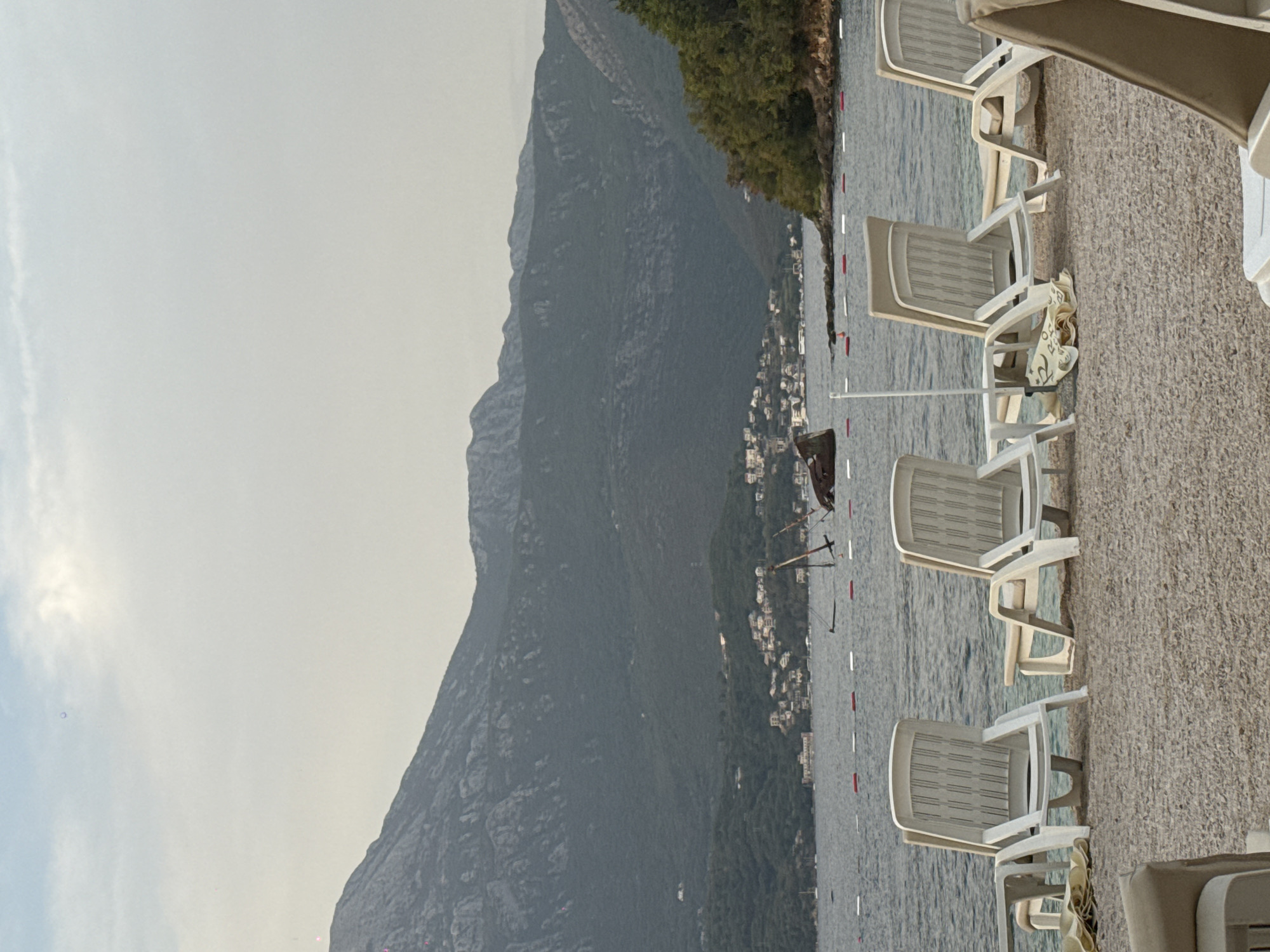 An outdoor, eye-level, vertical shot captures a beach with white lounge chairs in the foreground. The chairs are arranged on a pebble beach, with some towels and beach umbrellas visible. In the midground, calm water stretches out, marked by a line of red and white buoys. A partially submerged shipwreck is visible in the water, with masts sticking out. The background features a densely forested mountain range with a small town nestled at its base, and a hazy sky above. The overall scene evokes a tranquil, yet slightly melancholic, atmosphere.