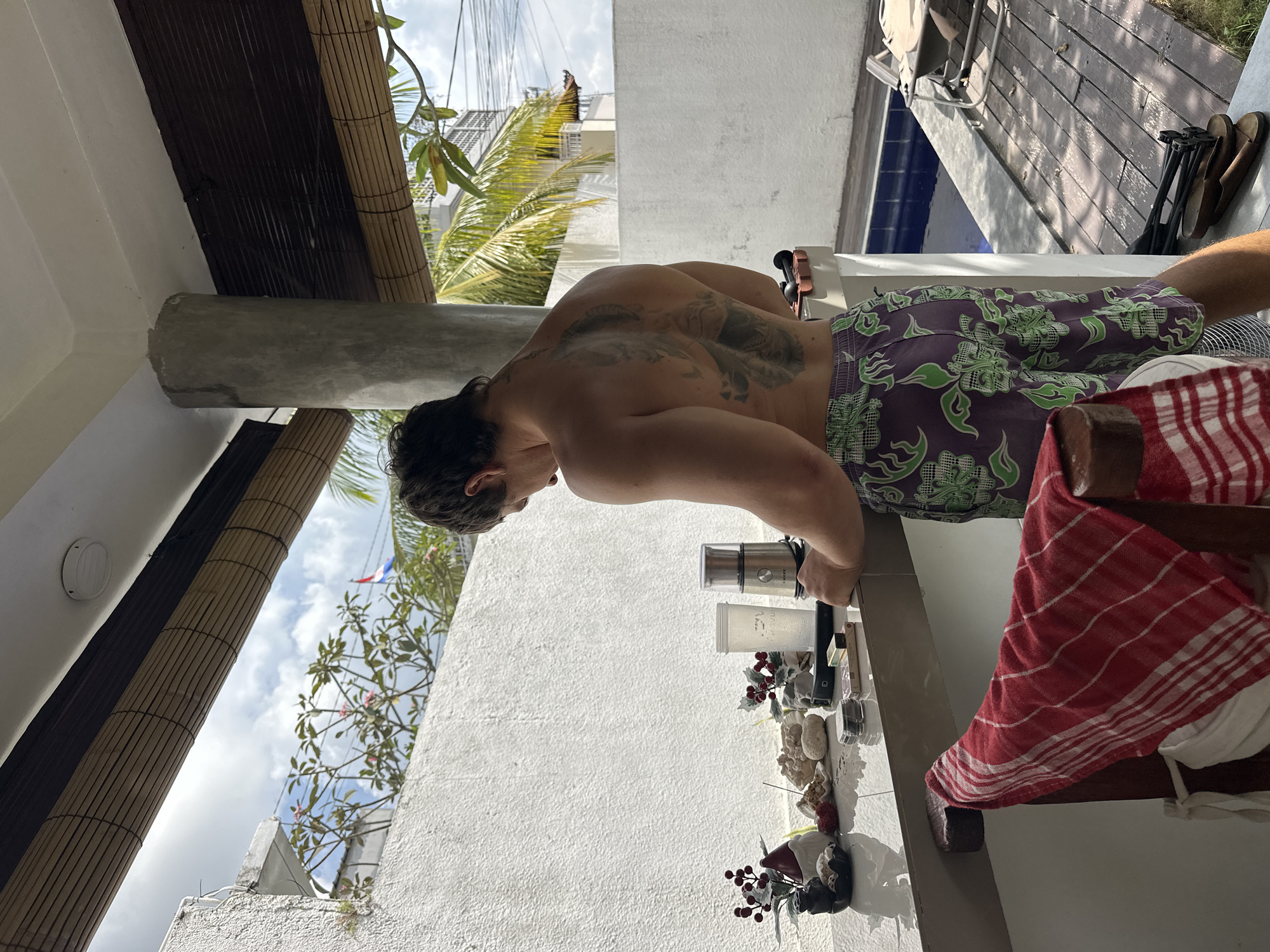 A shirtless man with a large tattoo on his back stands in front of a counter, looking down at items on the surface. He is wearing purple patterned shorts. On the counter are a small ceramic gnome with red berries, some stones, and a coffee grinder with a cup. A red and white striped towel hangs over a chair in the foreground. In the background, a white wall, some greenery, and a swimming pool are visible. The scene appears to be an outdoor patio area in a tropical location, possibly in North Kuta, Indonesia, given the presence of lush vegetation and tropical architectural elements like the bamboo blinds.
