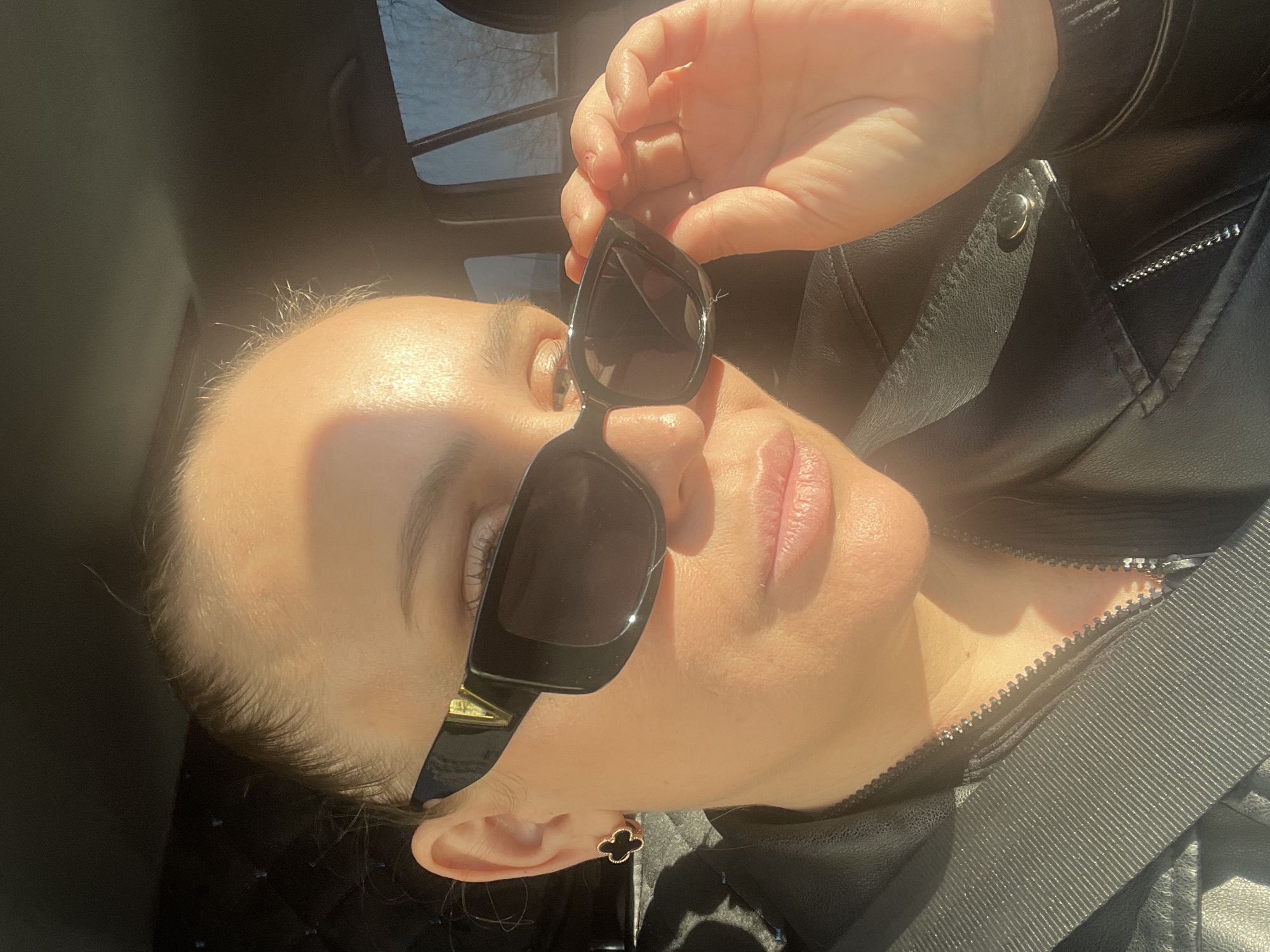 A person is in a vehicle, holding sunglasses and wearing a black leather jacket. A seatbelt is visible across the chest. The vehicle has a sunroof. The location is not identifiable as Chaikovsky, Russia, or any specific place.