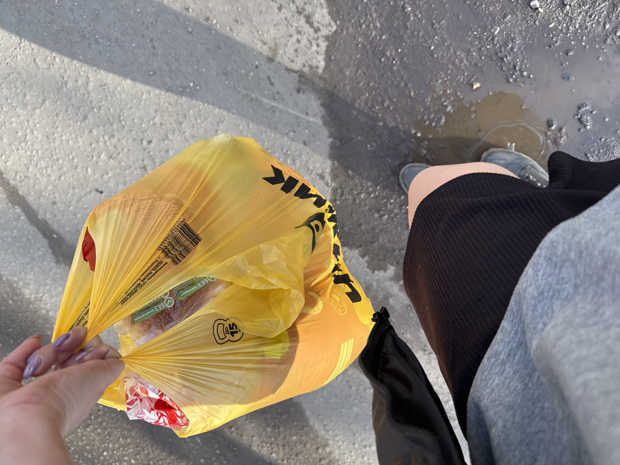A first-person view shows a person holding a yellow plastic bag from the "Dmitrovsky Molochny Zavod" (Dmitrov Dairy Plant) in Dmitrov, Russia. The bag contains food items, including bread and a package with red and white elements. The person is wearing a dark skirt or dress, a light gray sweater, and gray shoes. They are standing on a paved surface, possibly a sidewalk or road, with a puddle visible near their feet. Light and shadows suggest it is daytime. The hand holding the bag has long, light-colored fingernails.