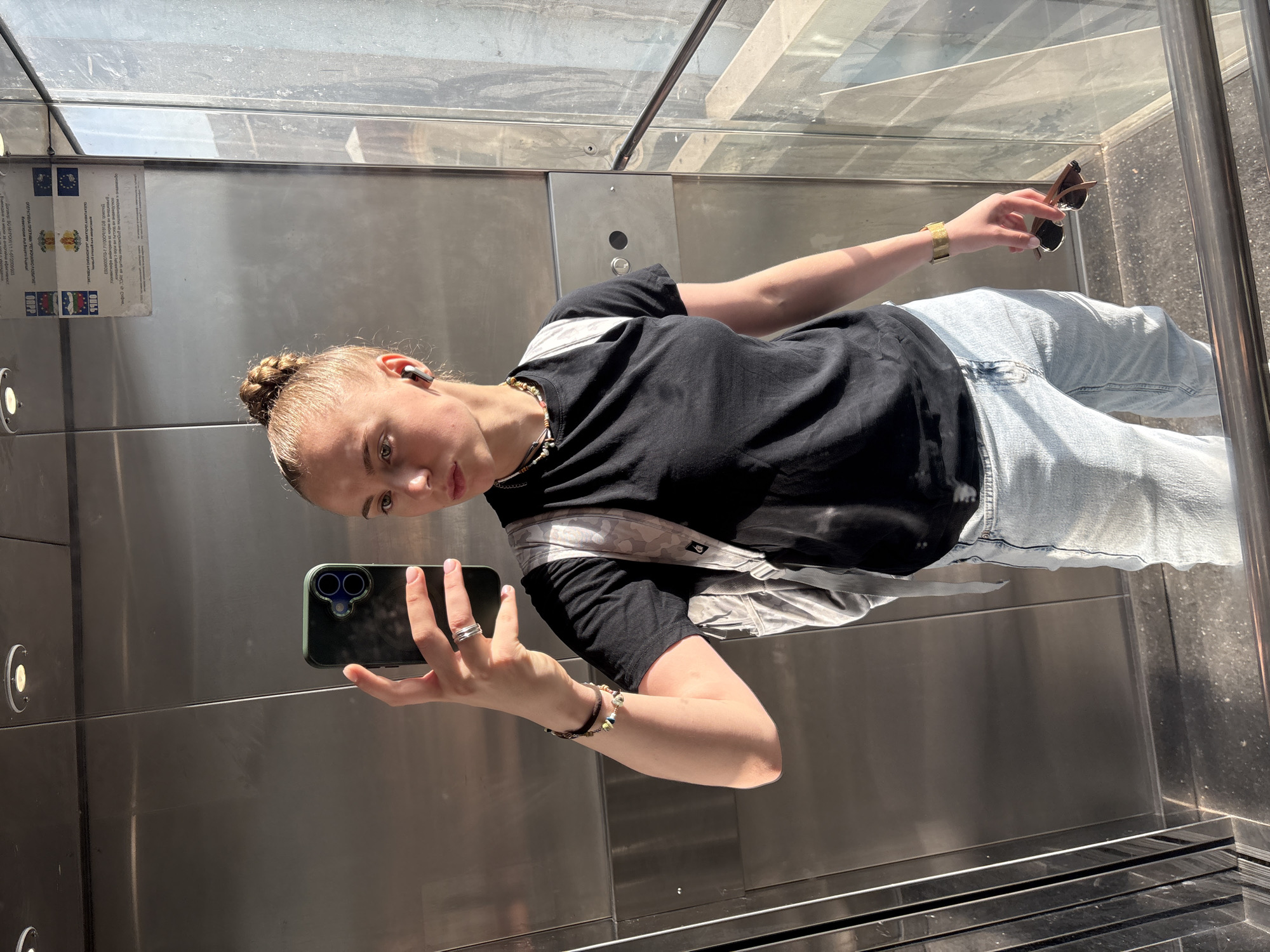 The image shows a young woman taking a selfie in what appears to be a modern, glass-walled elevator. She is wearing a black t-shirt, light blue jeans, and a crossbody bag. She is holding a smartphone in one hand and sunglasses in the other.
A sign is visible in the elevator, featuring the logos of the European Union and the Operational Programme "Regional Development" (OPRD) of Bulgaria. The sign also contains text related to a contract with ID number BG 161PO001/1.1-07/2008/001.
Based on the OPRD signage and the urban setting, it is likely that the photo was taken in Sofia, Bulgaria.