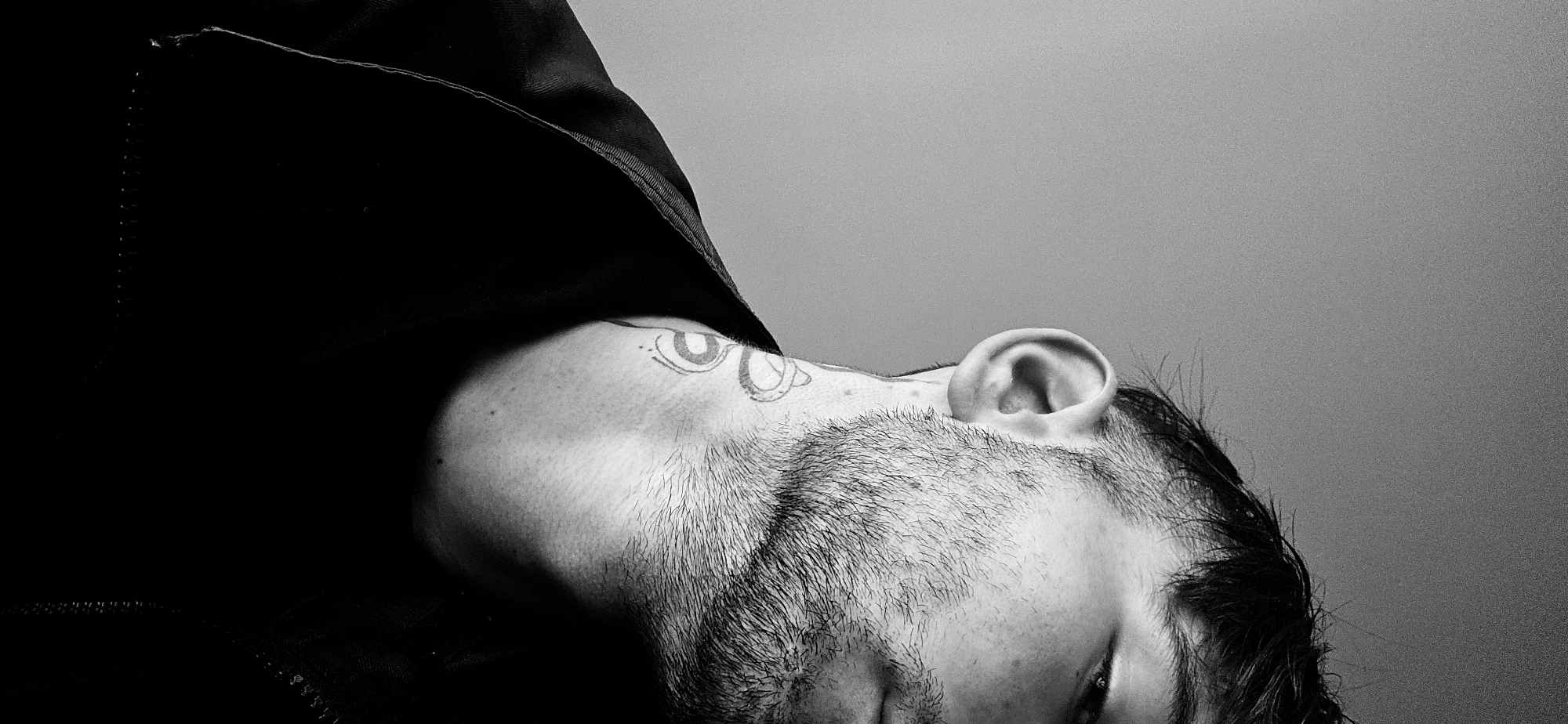A black and white close-up portrait captures a person, likely male, with their head tilted dramatically upwards and to the left. The focus is on the right side of their face, neck, and ear, against a plain, light gray background. The individual has short facial hair, appearing as stubble or a short beard, and dark hair that is partly visible near the ear. A distinctive tattoo featuring abstract, circular designs is visible on their left neck/shoulder area.
A dark piece of clothing, possibly a hood or collar, covers the upper left portion of the frame, creating a deep shadow that contrasts with the illuminated skin. The person's gaze is directed upwards, with their eyes not fully open, conveying a sense of introspection or contemplation. The strong, high-contrast lighting highlights the textures of the skin and hair, and sculpts the contours of the face. There are no overt activities taking place, rather a deliberate pose for a portrait. No text is visible within the image. The setting appears to be an indoor, controlled environment, possibly in Plovdiv, Bulgaria.
