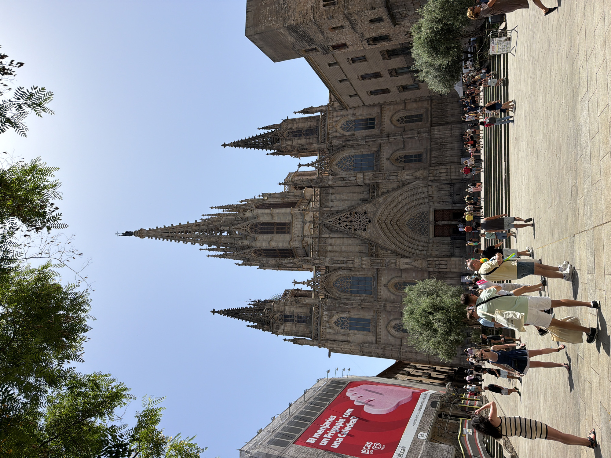 This is a vertical, eye-level, outdoor shot of the Barcelona Cathedral on a sunny day. The cathedral, made of stone, has intricate gothic architecture with spires and arched windows. In the foreground, a group of people are walking on a paved square, some looking up at the cathedral. The sun casts long shadows on the ground, indicating it might be late afternoon. A large red banner with text in Catalan is draped over scaffolding on the right side of the frame. The text on the banner reads, "El menjaries un Frigopie com una Catedral?" which translates to "Would you eat a Frigopie like a Cathedral?". This appears to be an advertisement. The sky is clear and blue. The scene evokes a sense of tourism and everyday life in a historic city.